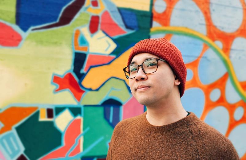 Mann med briller, brun strikkegenser og rød lue poserer framfor fargerik, tagget betongvegg.