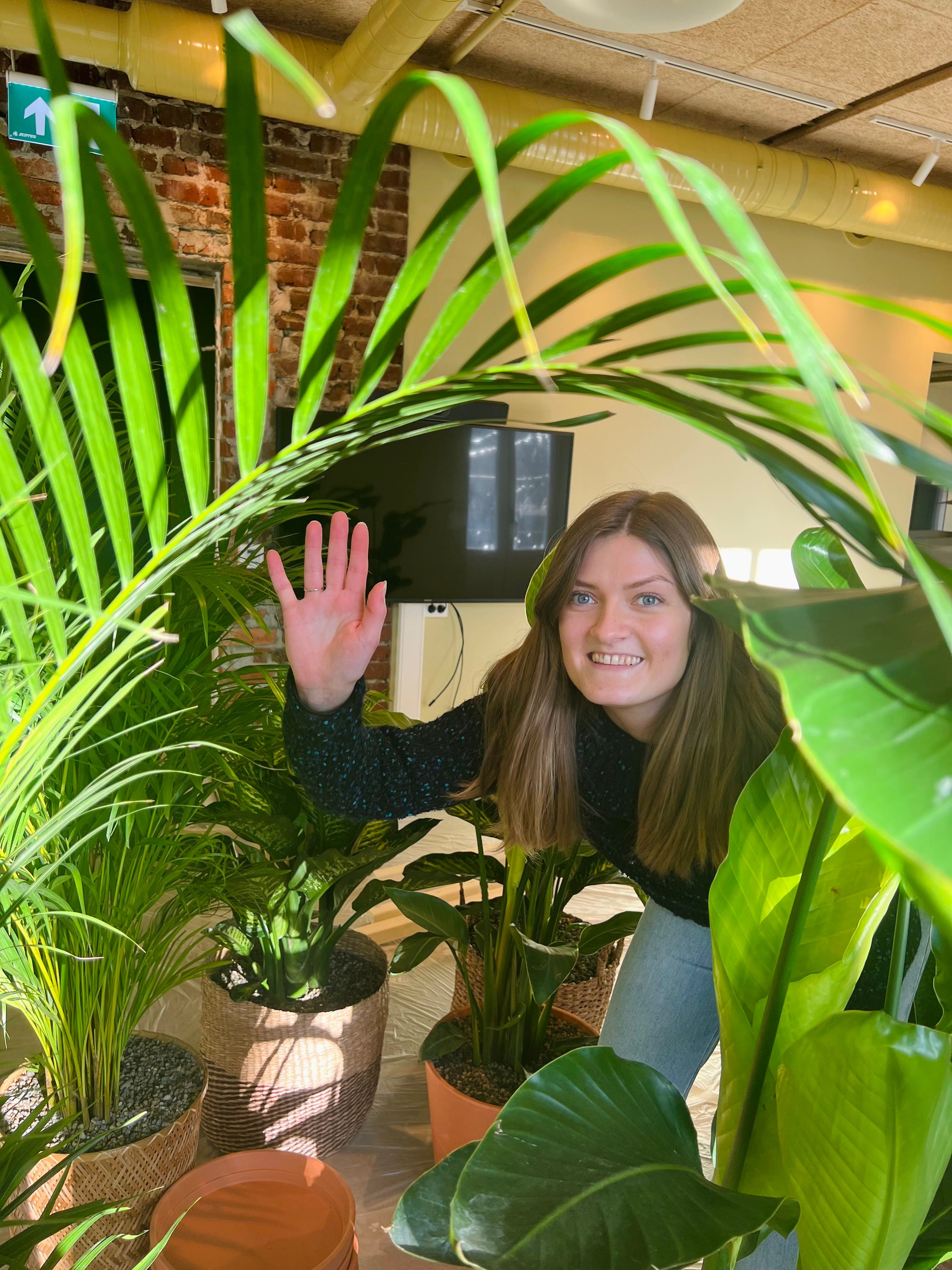 Dame med langt hår vinker til kamera gjennom flere grønne potteplanter.