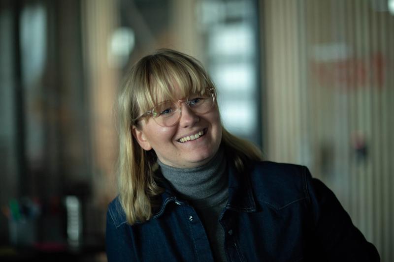 Dame med lyst hår, lugg og briller. Hun går med dongeri-jakke og grå turtle-neck genser.