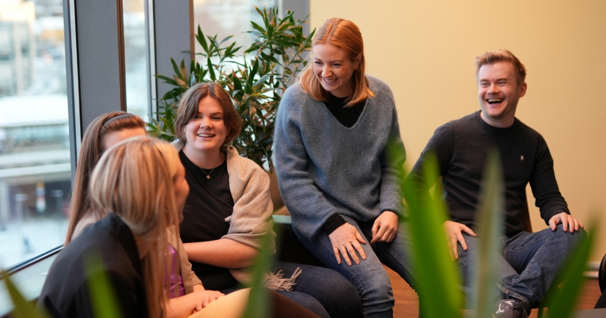 5 UX designere sitter sammen og snakker til hverandre. Alle smiler og ler. De sitter i et gult rom med et stort video bak dem.