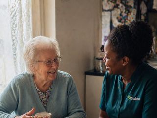 An Oakma carer chatting with an older woman over a pot of tea at her kitchen table