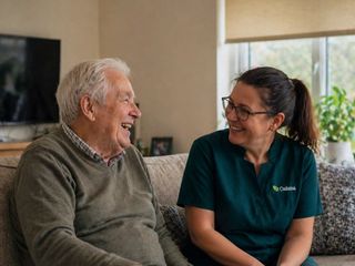 An Oakma carer and an older man laughing together on the sofa in his living room
