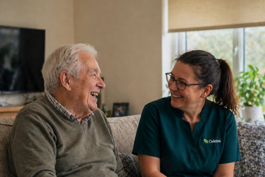 An Oakma carer and an older man laughing together on the sofa in his living room