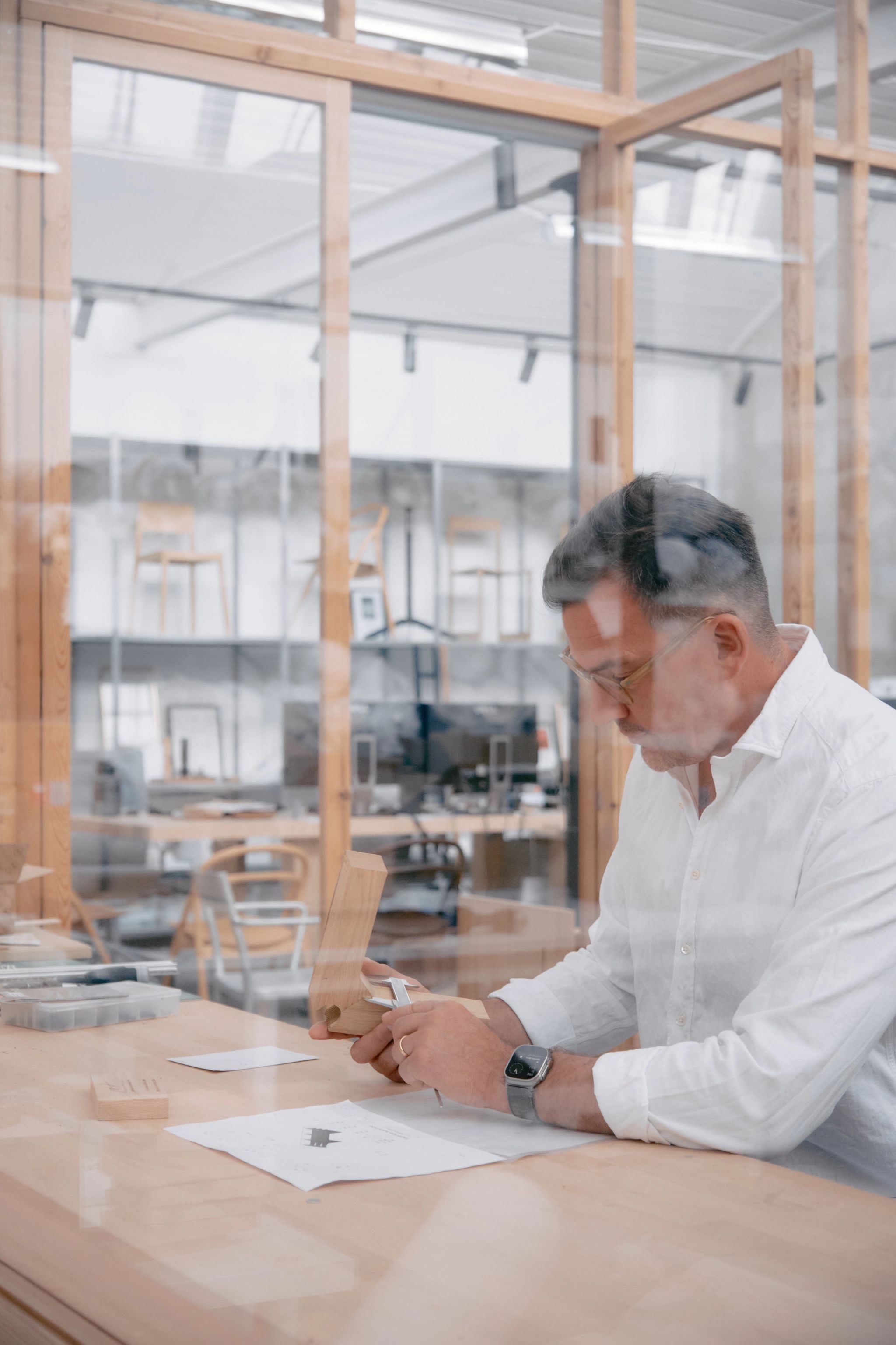 Designer Staffan Holm measuring a wooden prototype in a bright, modern studio with glass walls and wooden furniture.