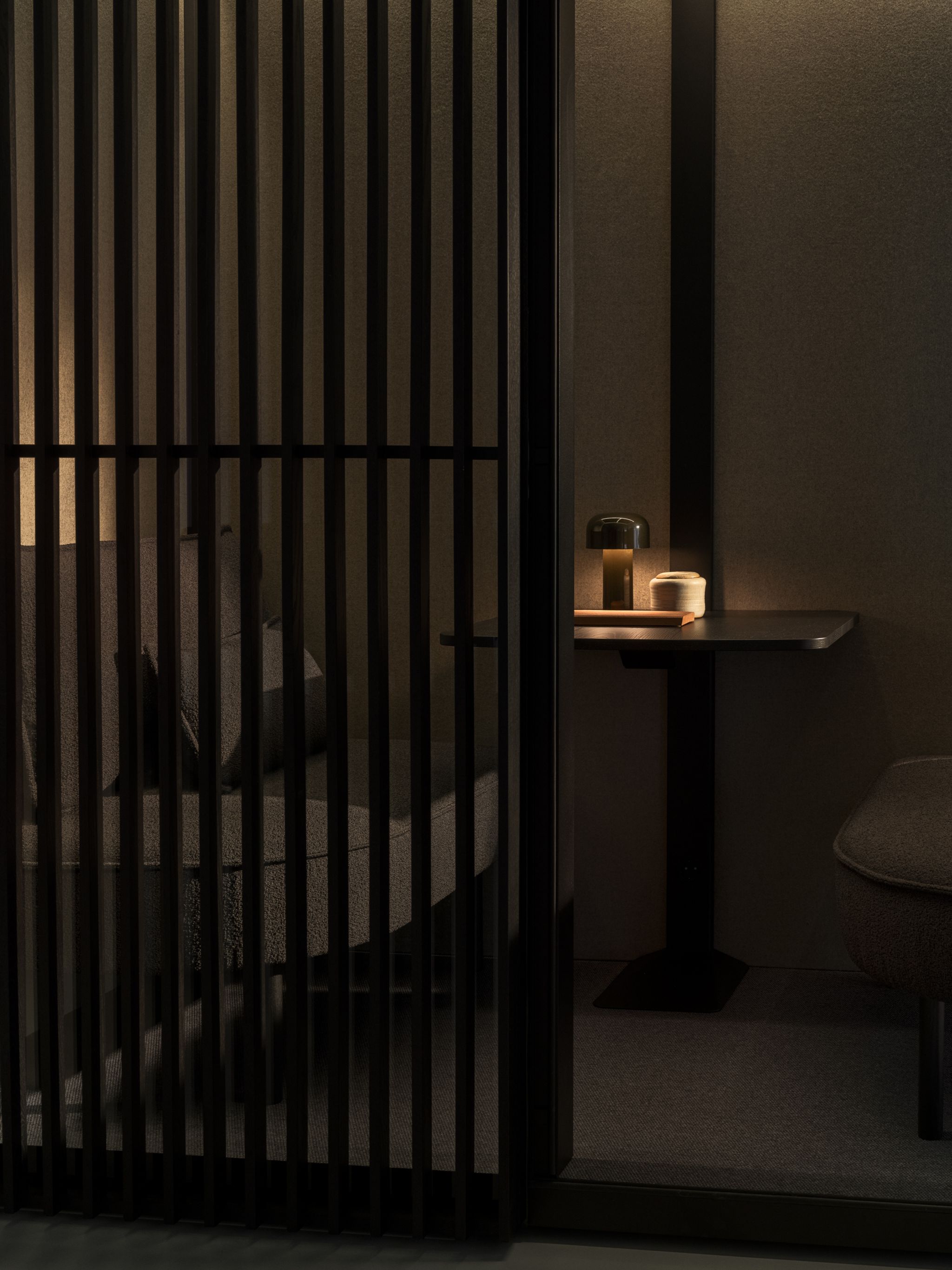 Dimly lit Zen Pod workspace with a small table, lamp, and cushioned seating behind black vertical slats.