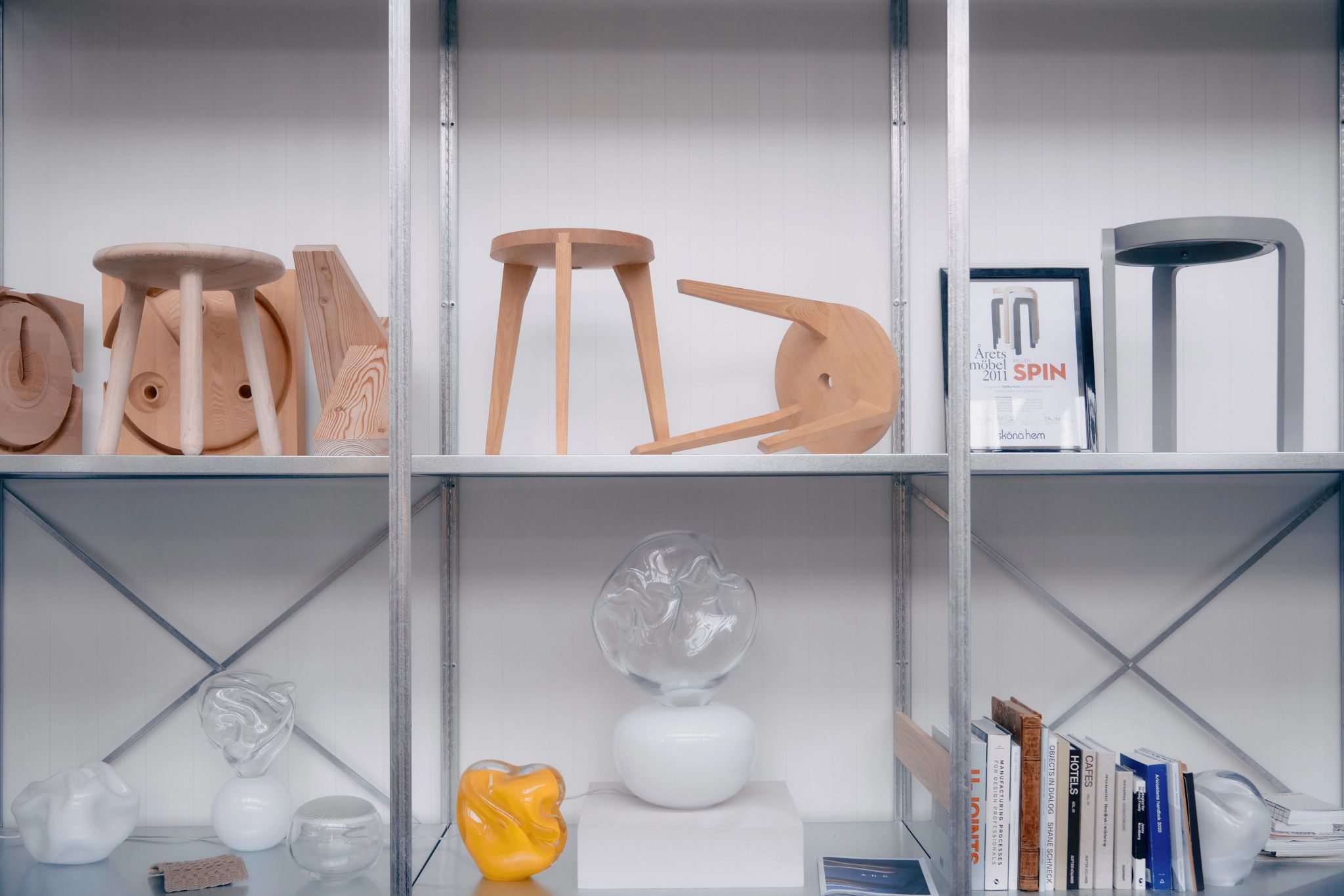 Metal shelving unit displaying wooden stools, glass sculptures, books, and a framed design award certificate.