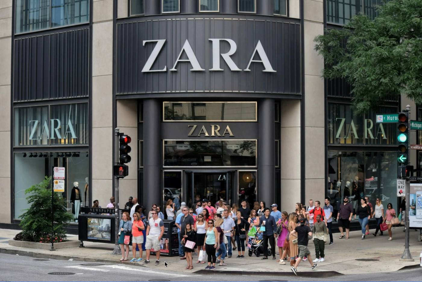 Busy Zara storefront on a city street with crowds of shoppers gathered outside the large black and beige facade, multiple windows displaying minimalist product displays