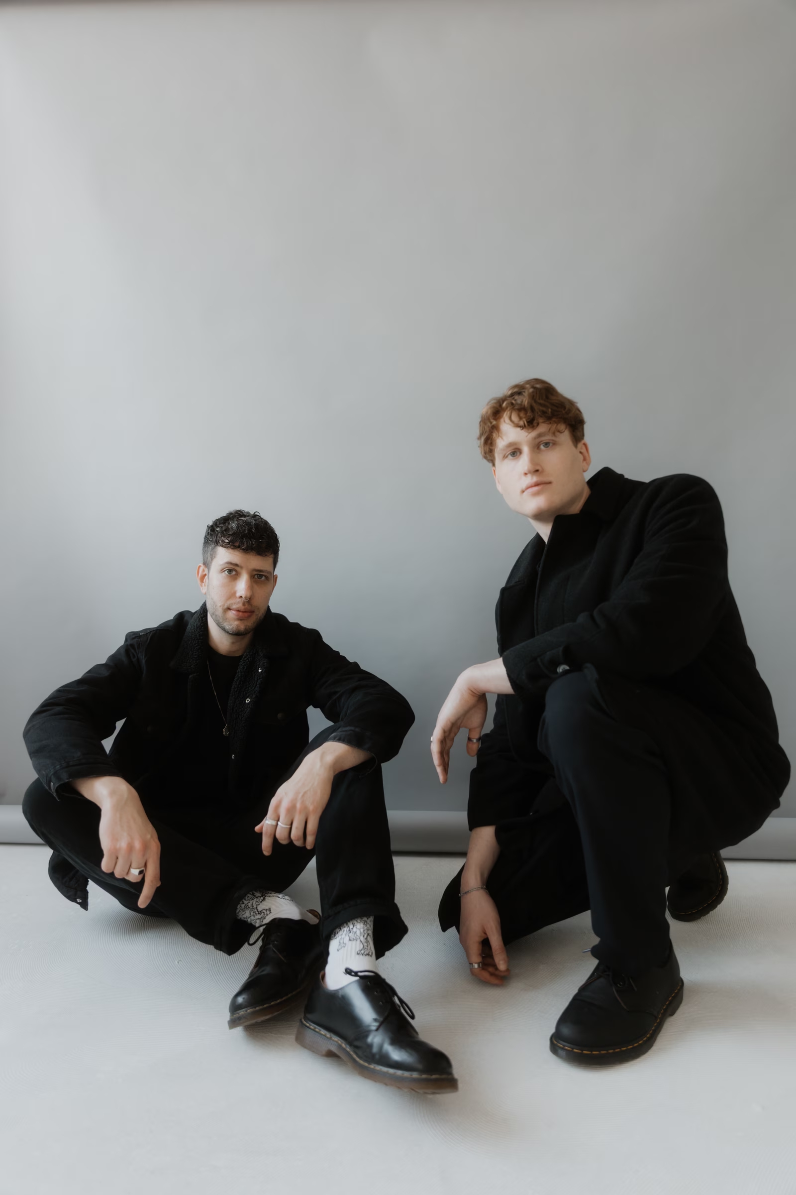 Adrian and Julian dressed in black sitting on the floor against a neutral studio wall, posed casually in a minimalist setting