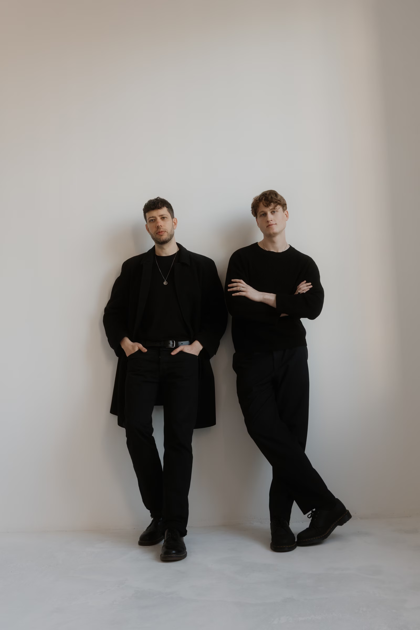Adrian and Julian dressed in black standing against a white studio wall, posed casually in a minimalist setting