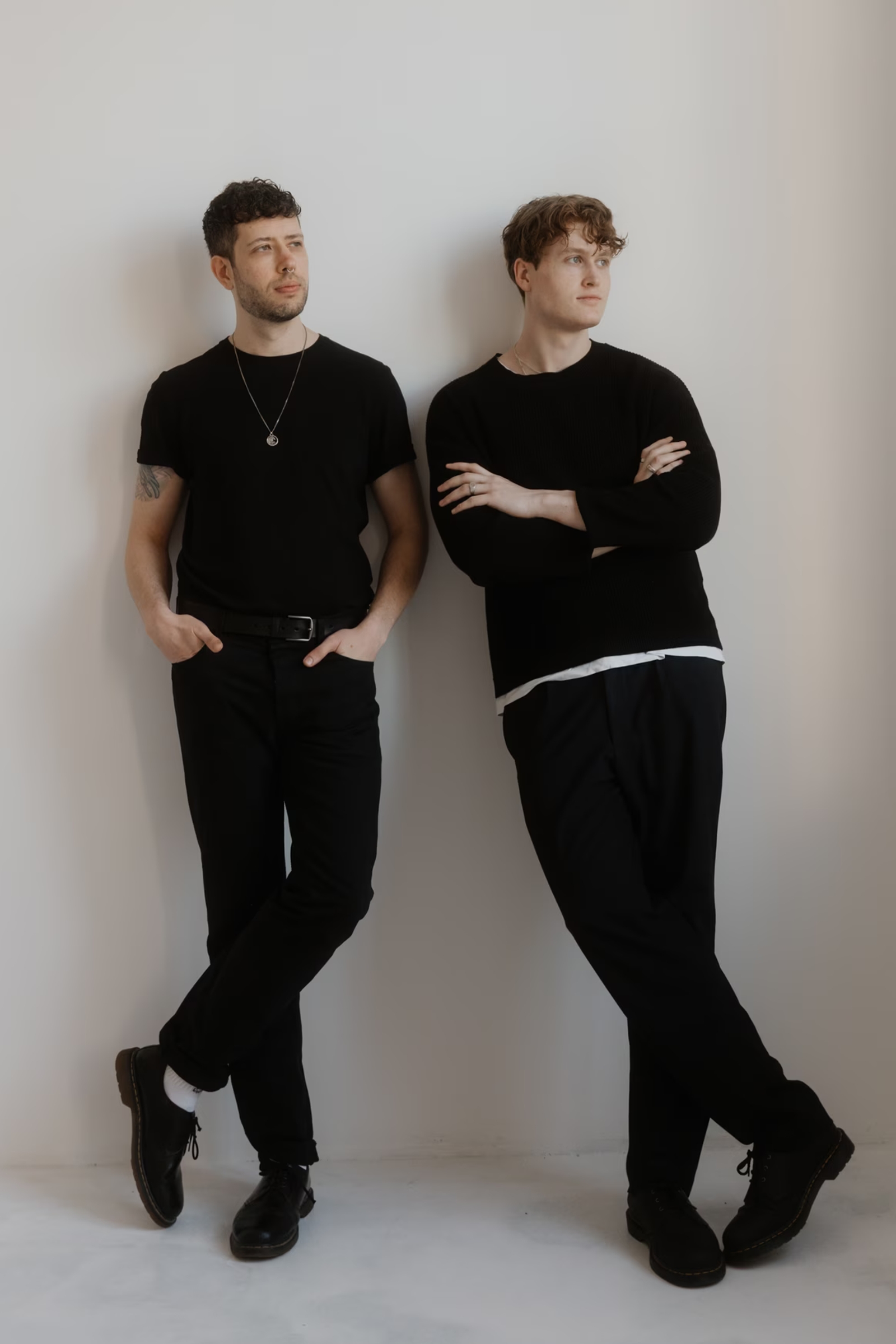 Adrian and Julian dressed in black standing against a white wall, looking off to the side in a minimalist studio