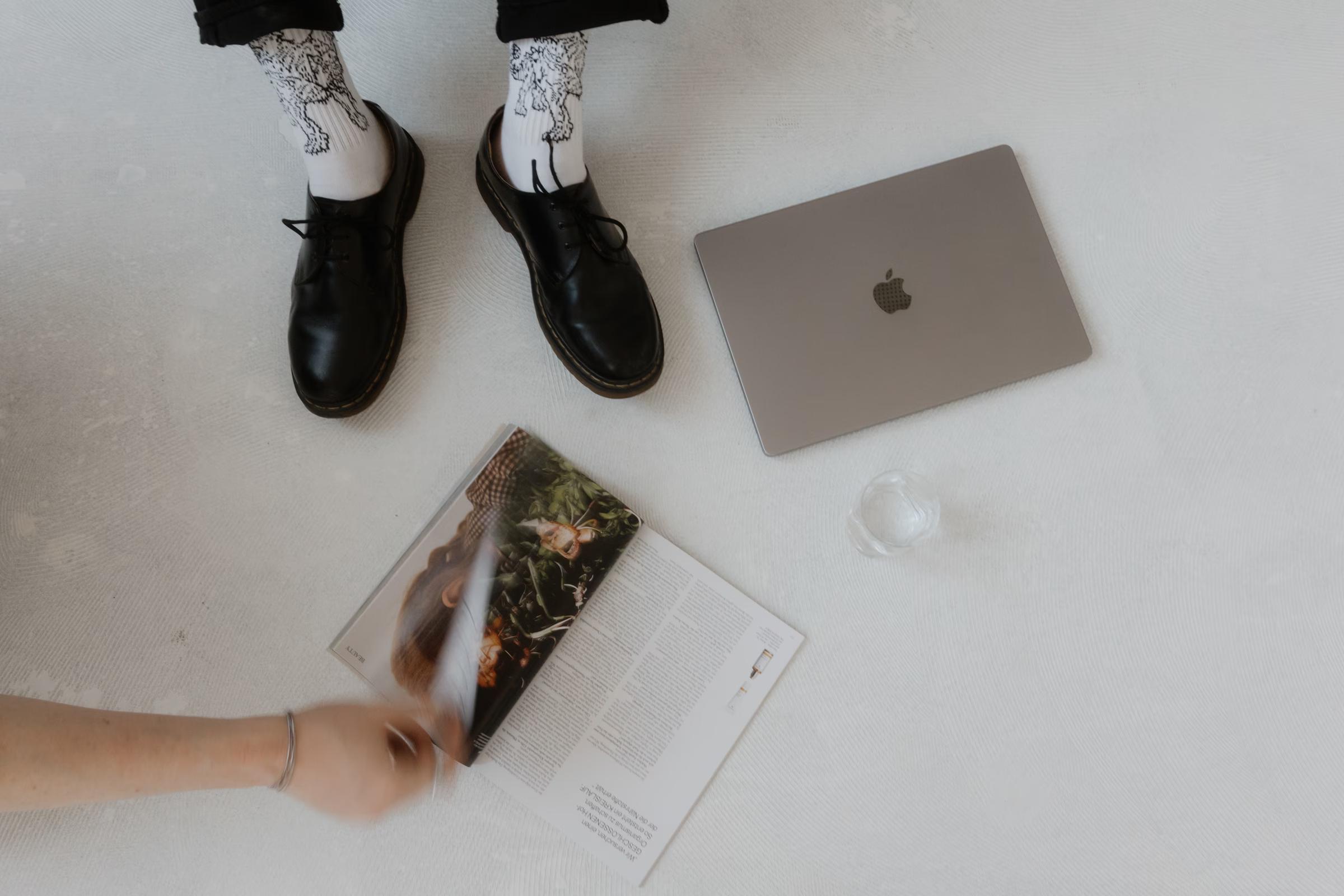 Top-down view of black shoes and patterned socks on a white floor beside an open magazine, a closed laptop, and a glass of water