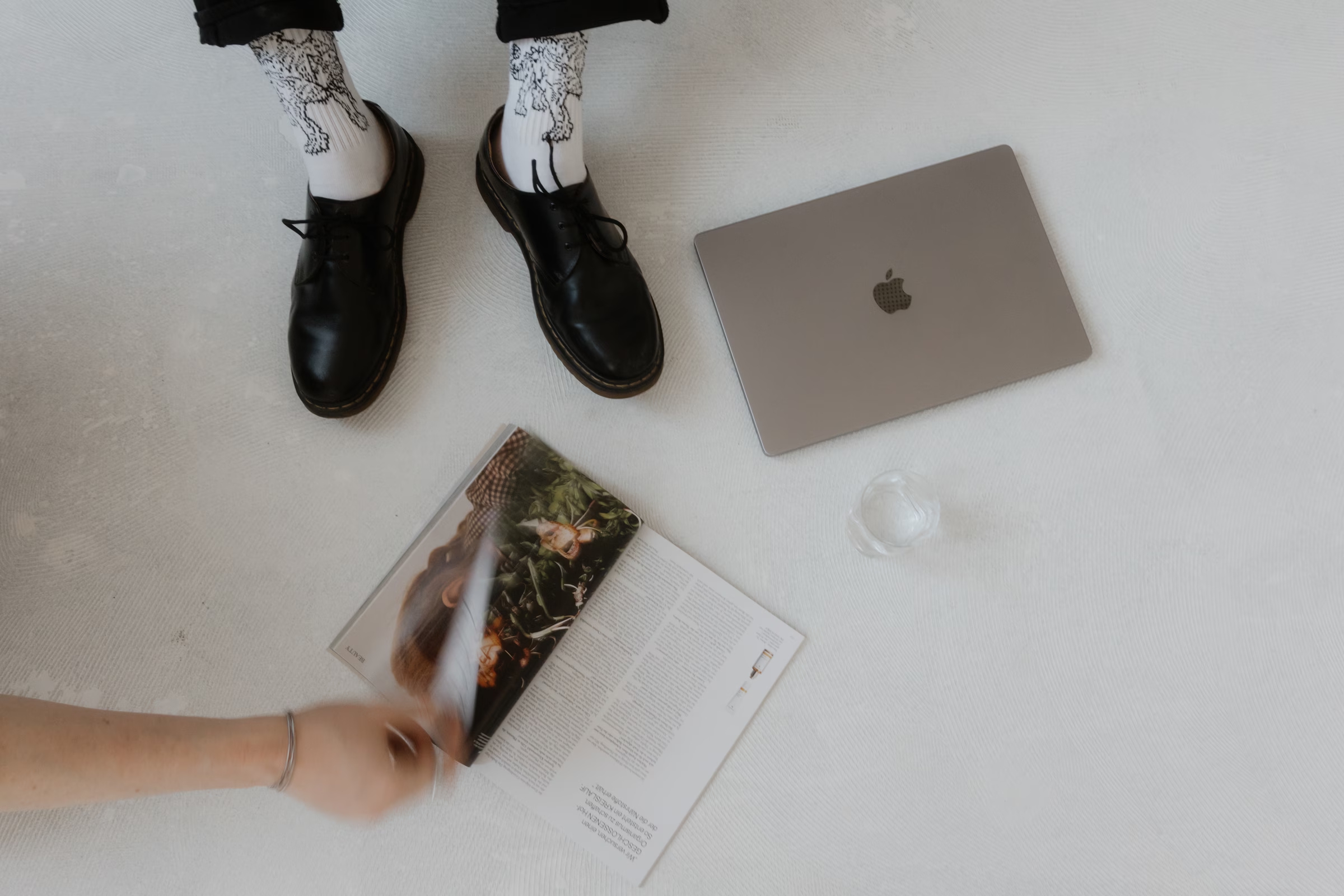 Top-down view of black shoes and patterned socks on a white floor beside an open magazine, a closed laptop, and a glass of water