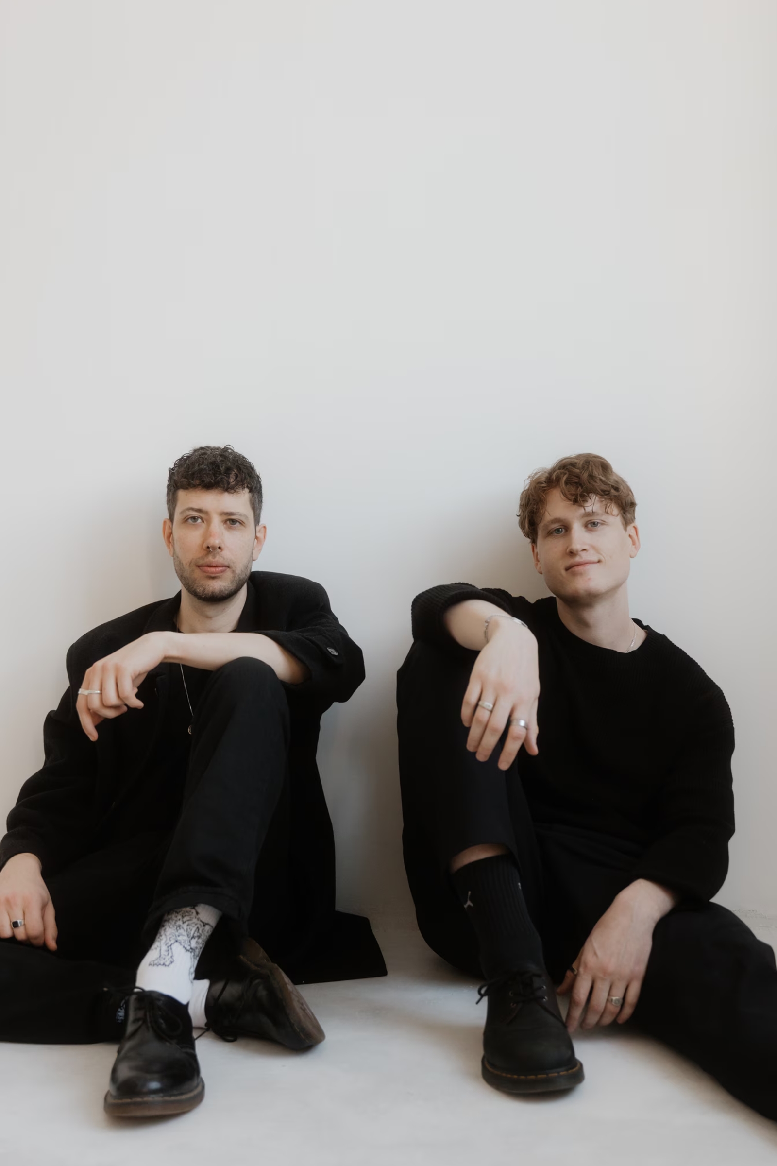 Adrian and Julian dressed in black sitting on the floor against a white wall in a minimalist studio setting