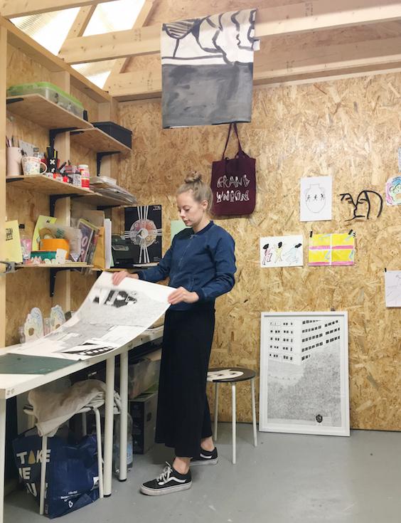 A woman stands holding some paper in an artist's studio