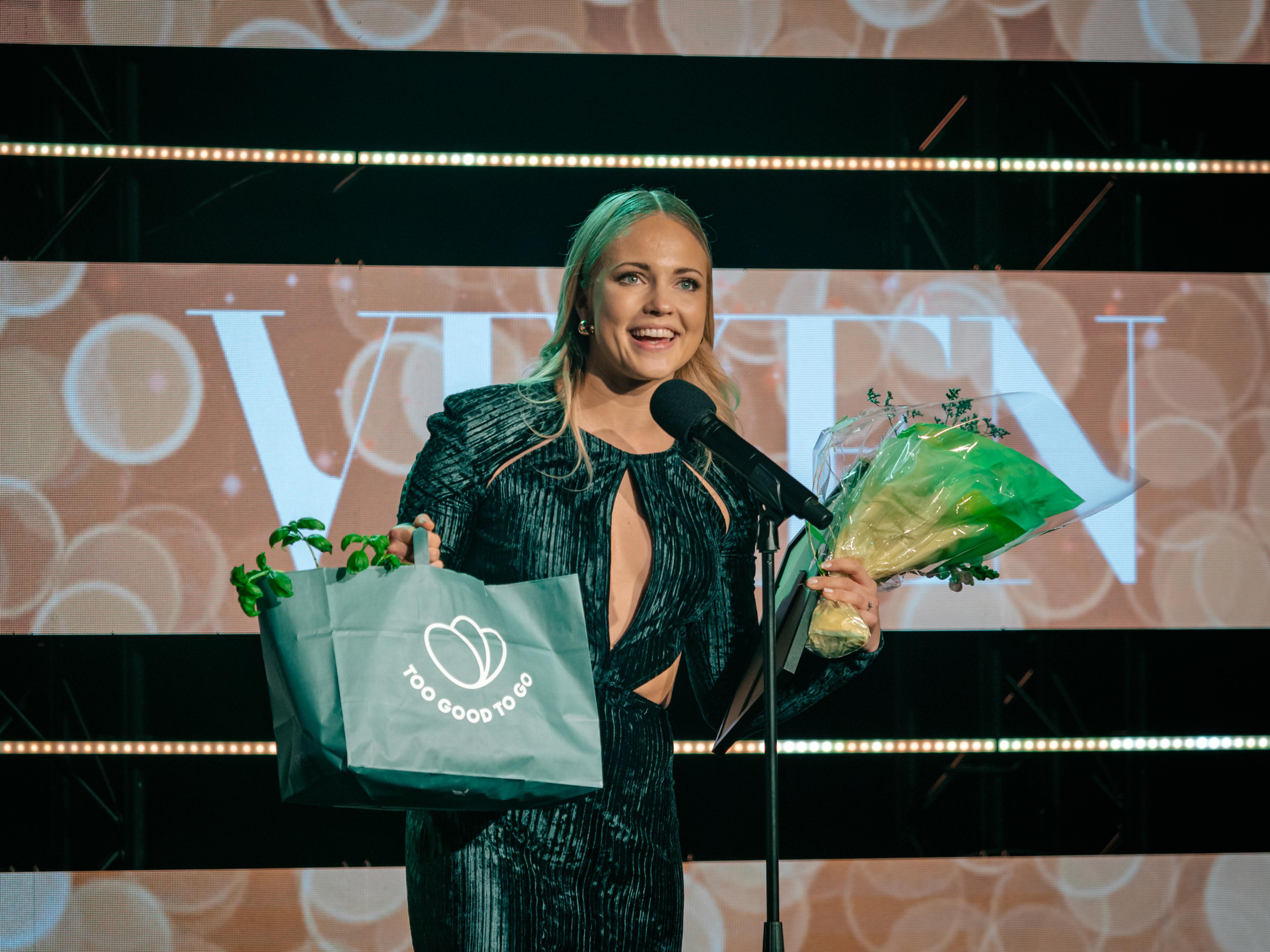 Emilie Nereng står på scenen og holder en pose fra Too Good To Go, og en blomsterbukett.