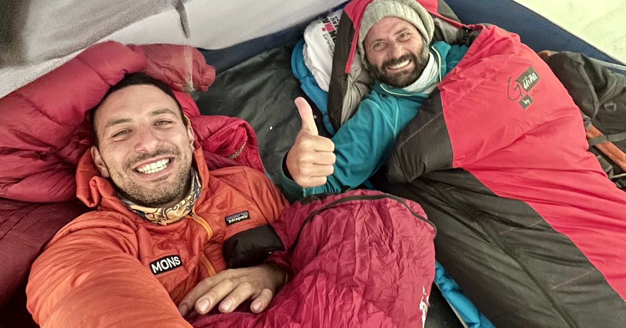 Dos trekkers de MONS sonriendo dentro de la carpa, abrigados en sus bolsas de dormir en un campamento de montaña