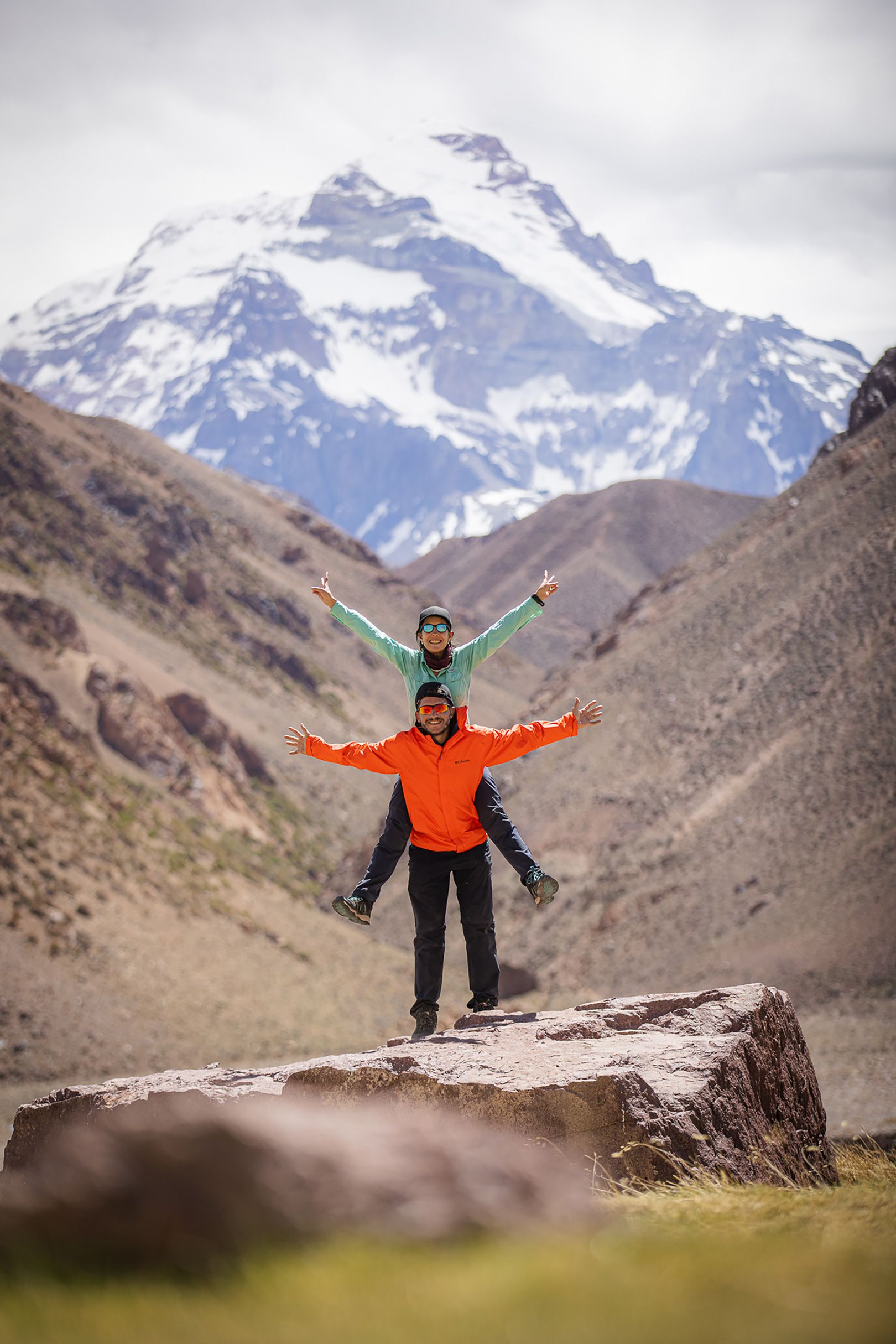 Dos pasajeros felices listos para recorrer la montaña, vistiendo ropa de montaña adecuada como camperas impermeables naranja y verde. De fondo, un paisaje de cerros y un gran pico nevado.