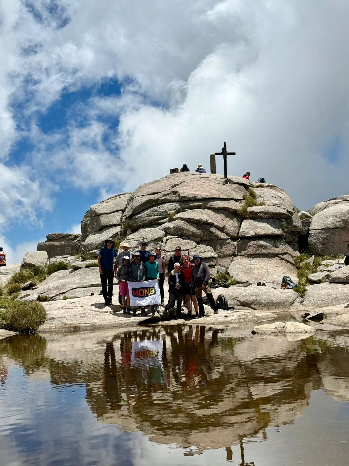 La cruz de la cumbre del Champaquí reflejada en la laguna con el grupo reunido sobre el granito