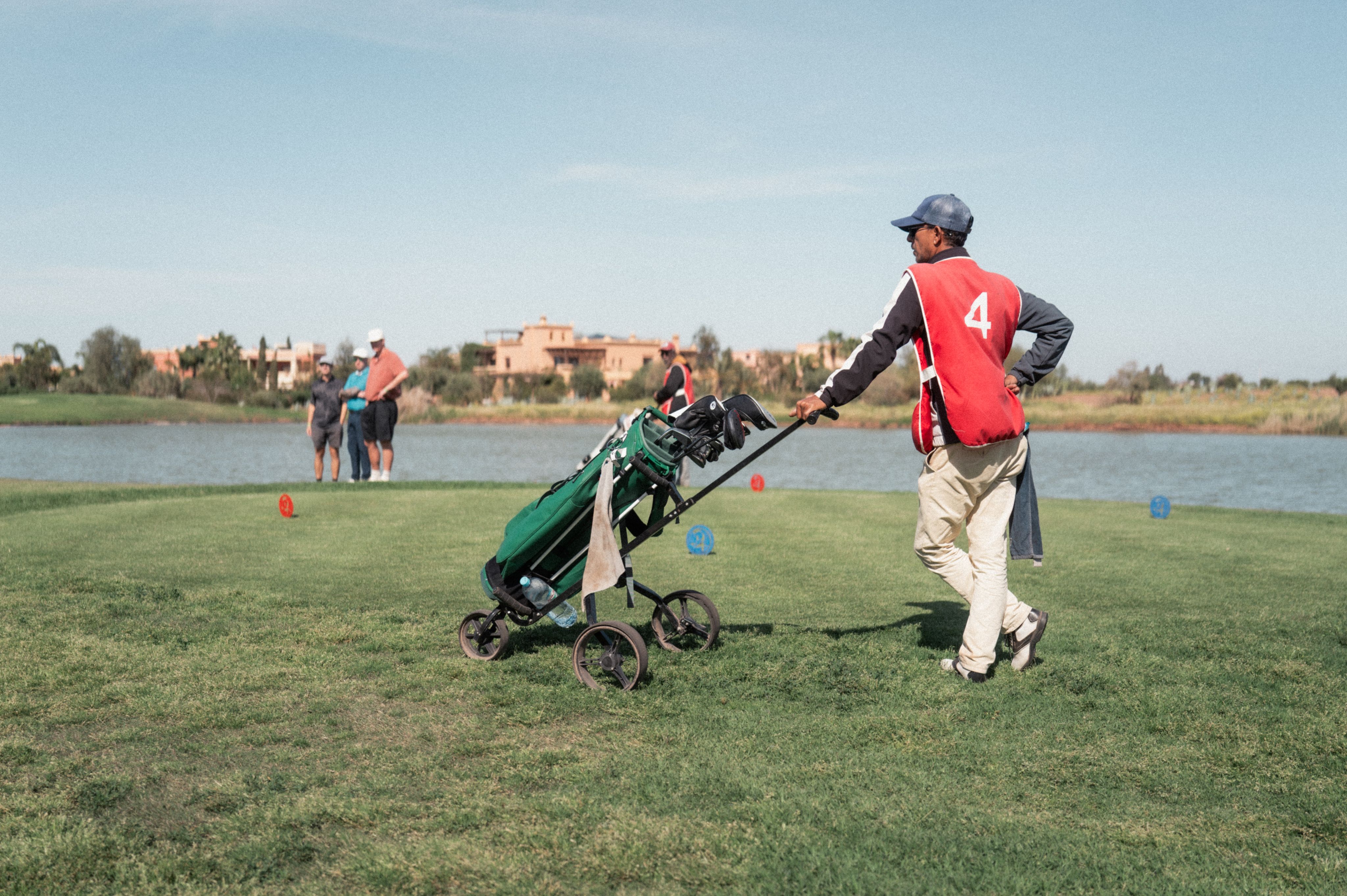 Caddie in a red bib pulling a golf trolley beside a lake on a sunny course in Morocco