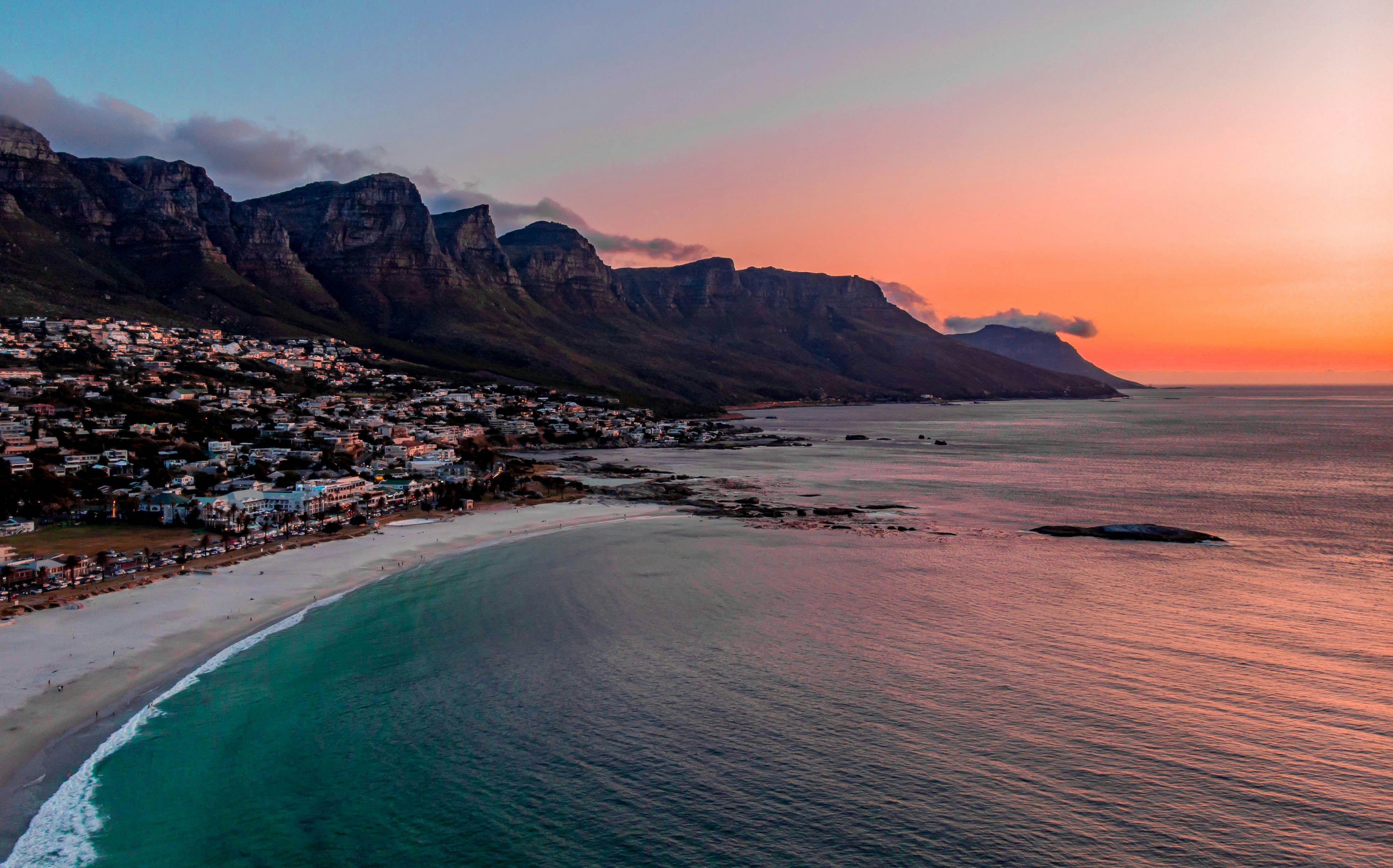 Coastal town beneath dramatic mountains at sunset, with a curving white-sand beach