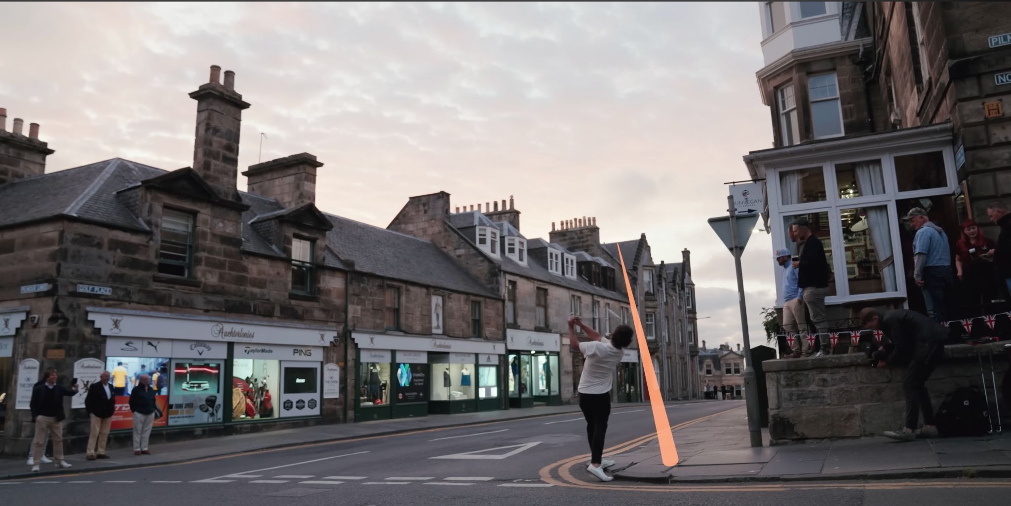 Golfer swinging in the street of a stone-built town at dusk as locals look on