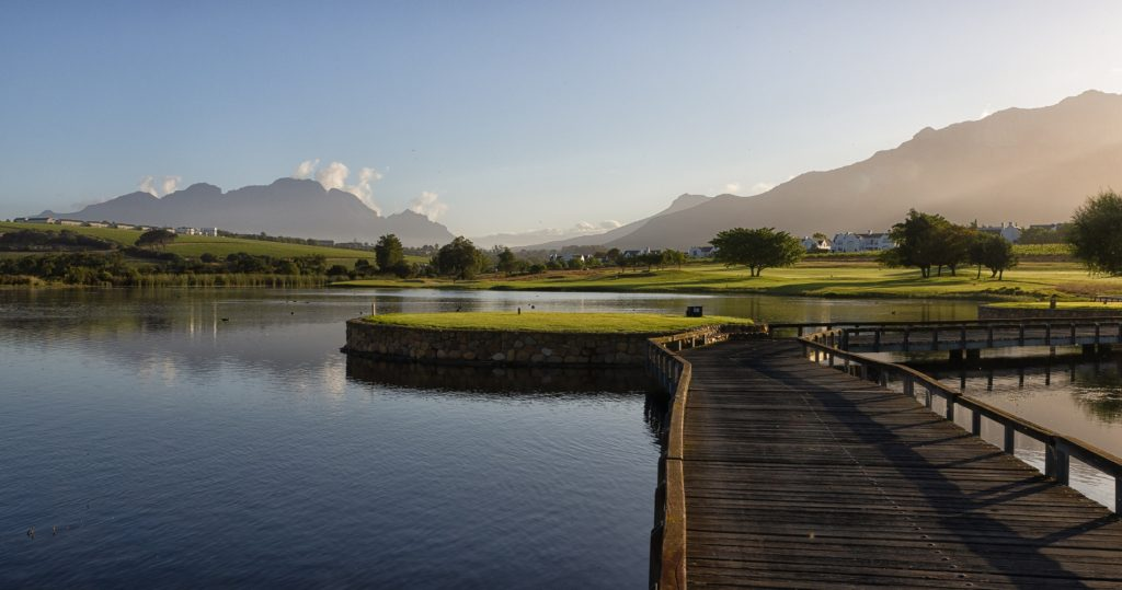 Wooden boardwalk crossing a calm lake toward a green, with mountains in the distance