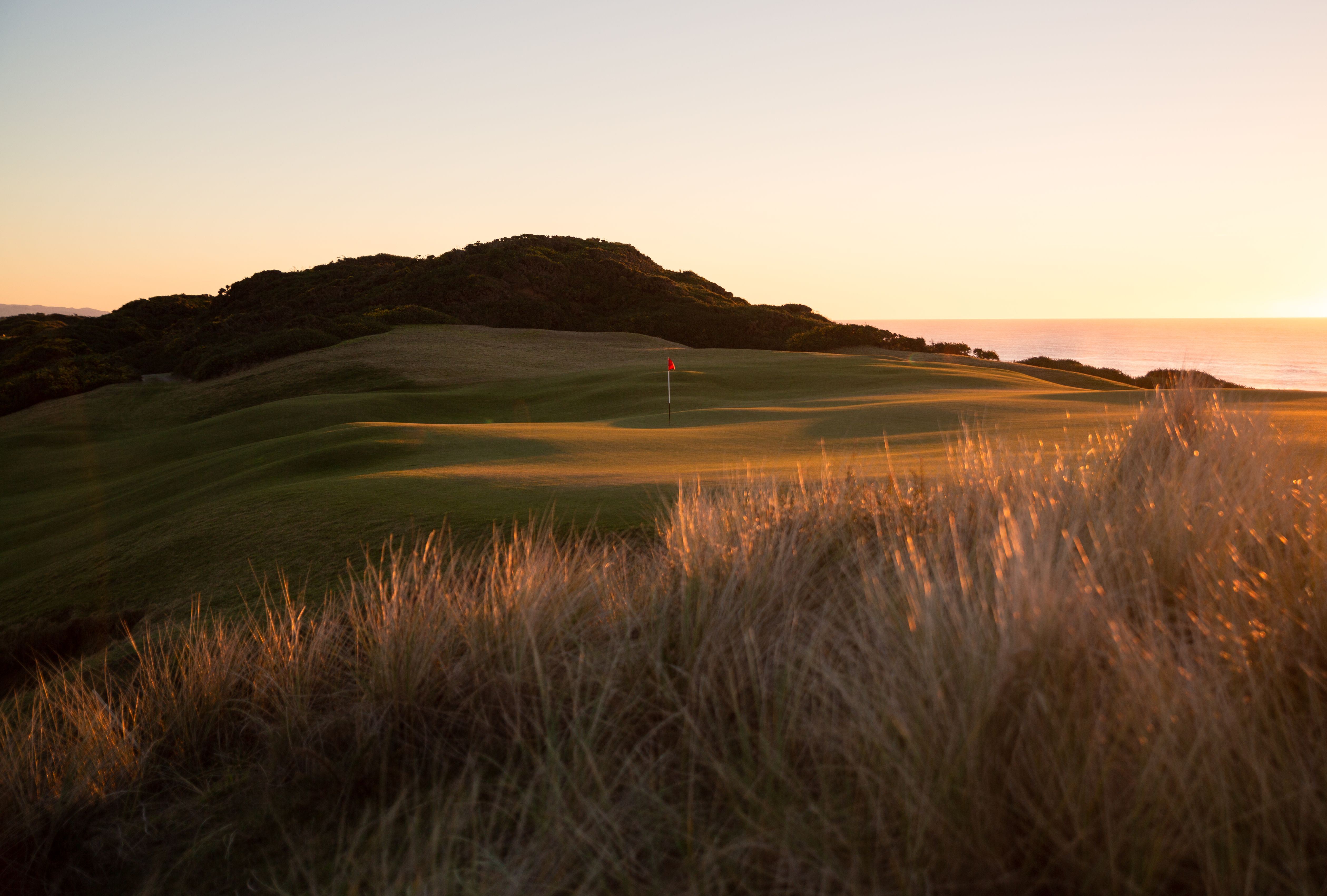 Old Macdonald golf course at sunset with a red flag on the green and ocean behind dune grass