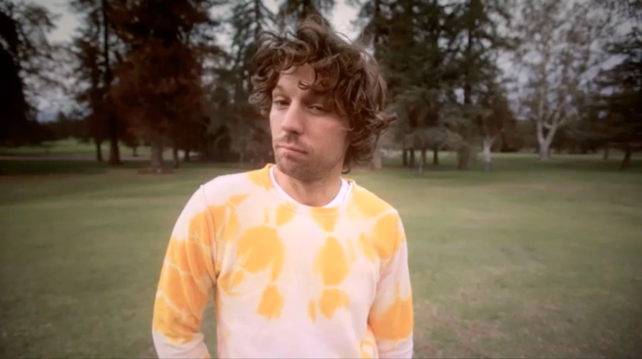 Curly-haired man in a tie-dye sweatshirt standing on a tree-lined golf course