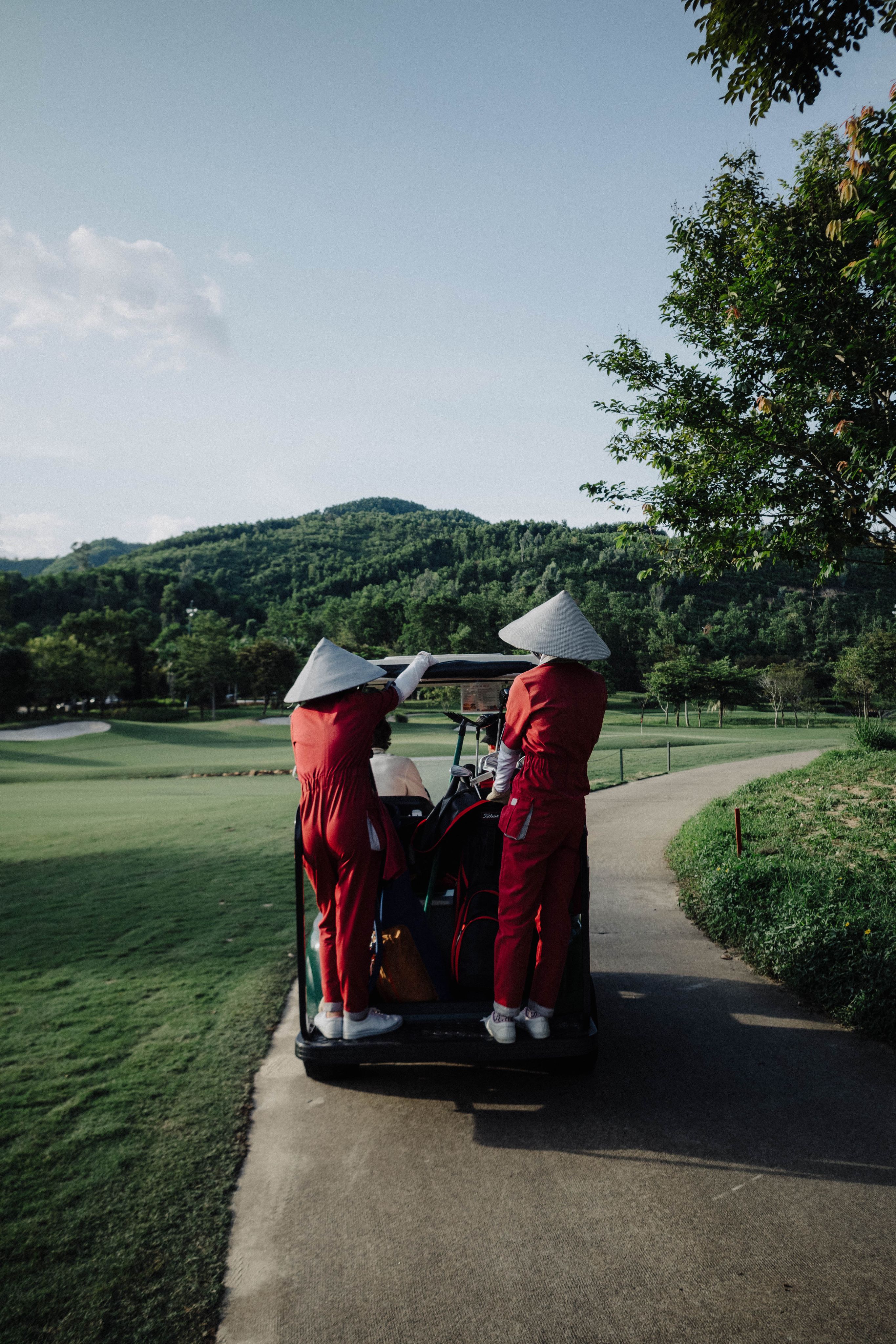 Caddies in red uniforms and conical hats riding the back of a golf cart, Vietnam