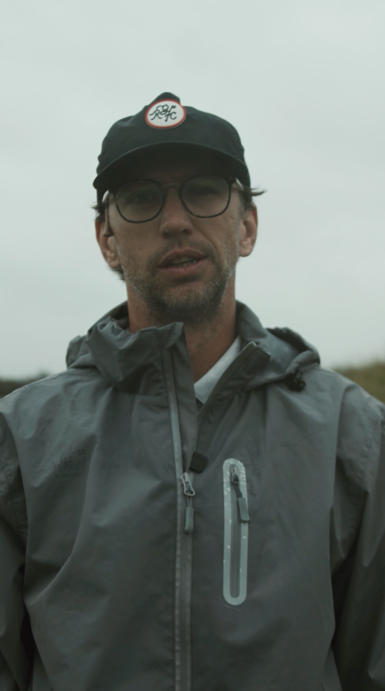 Man in a grey rain jacket and RGC cap speaking outdoors on an overcast day