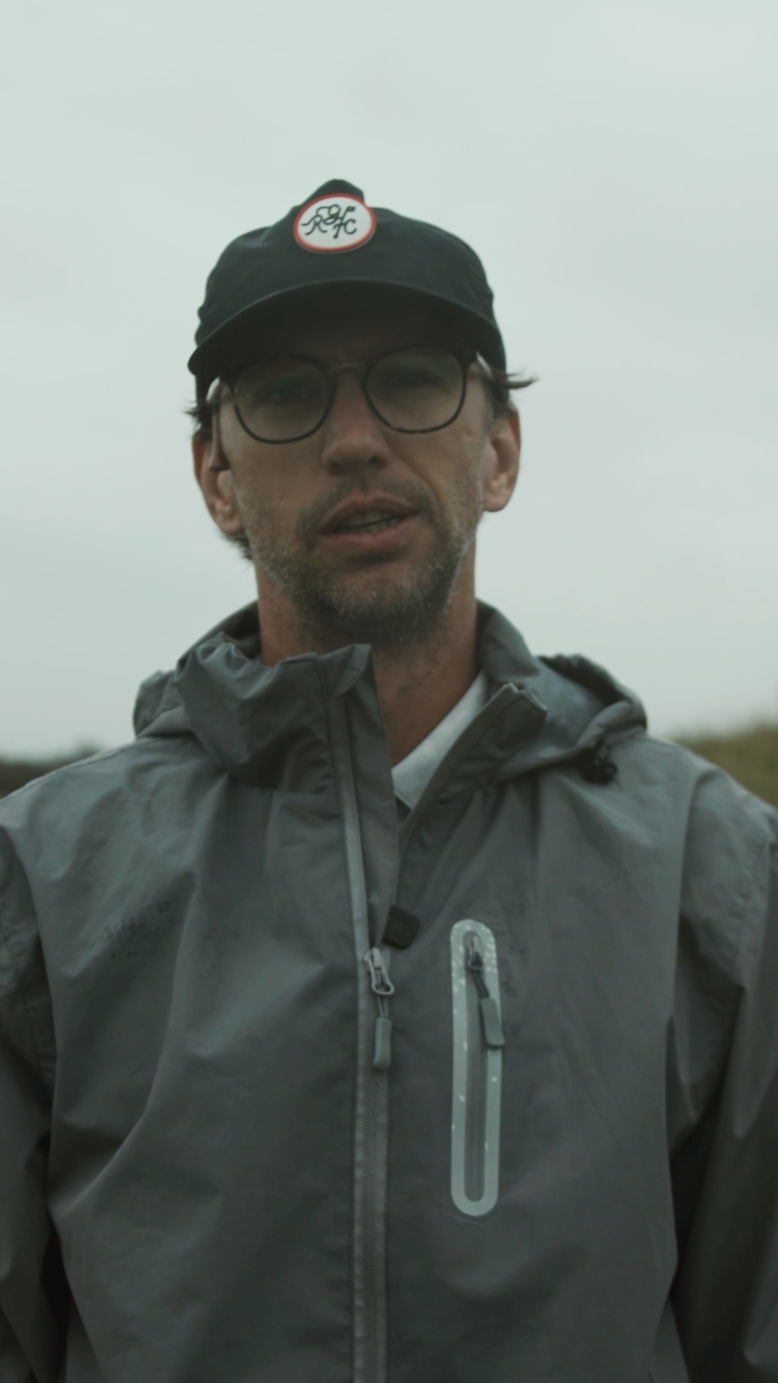 Man in a grey rain jacket and RGC cap speaking outdoors on an overcast day