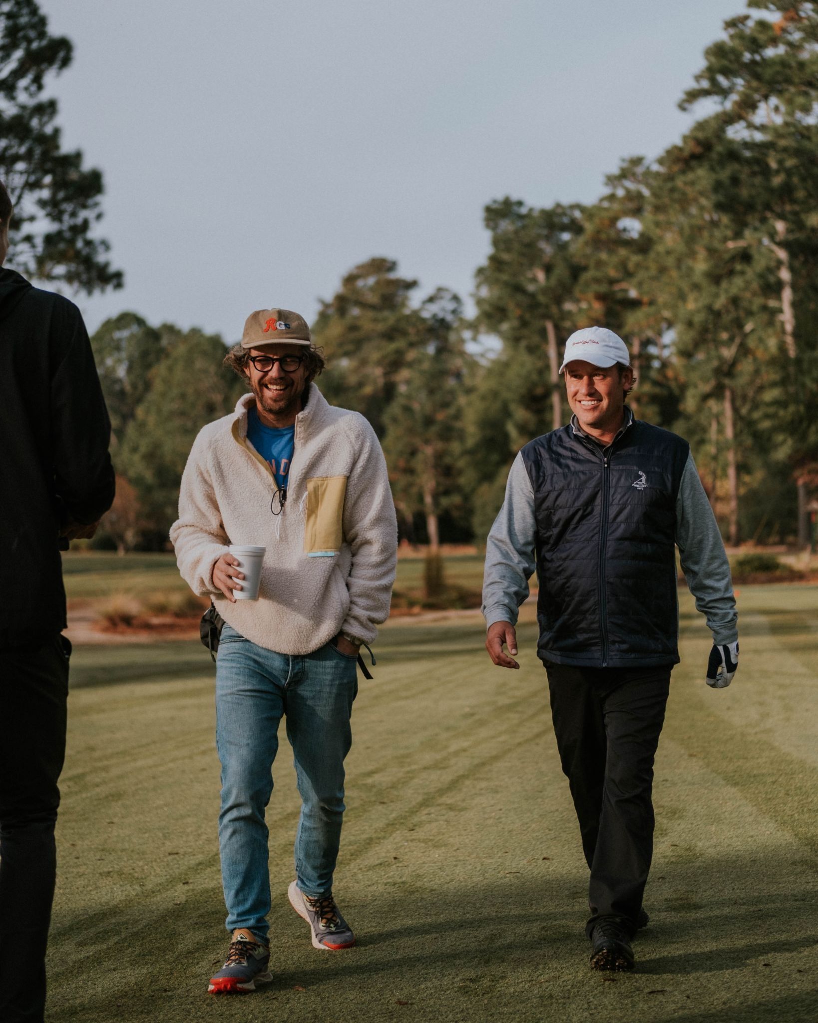 Two smiling men walking along a pine-lined fairway, one holding a coffee cup