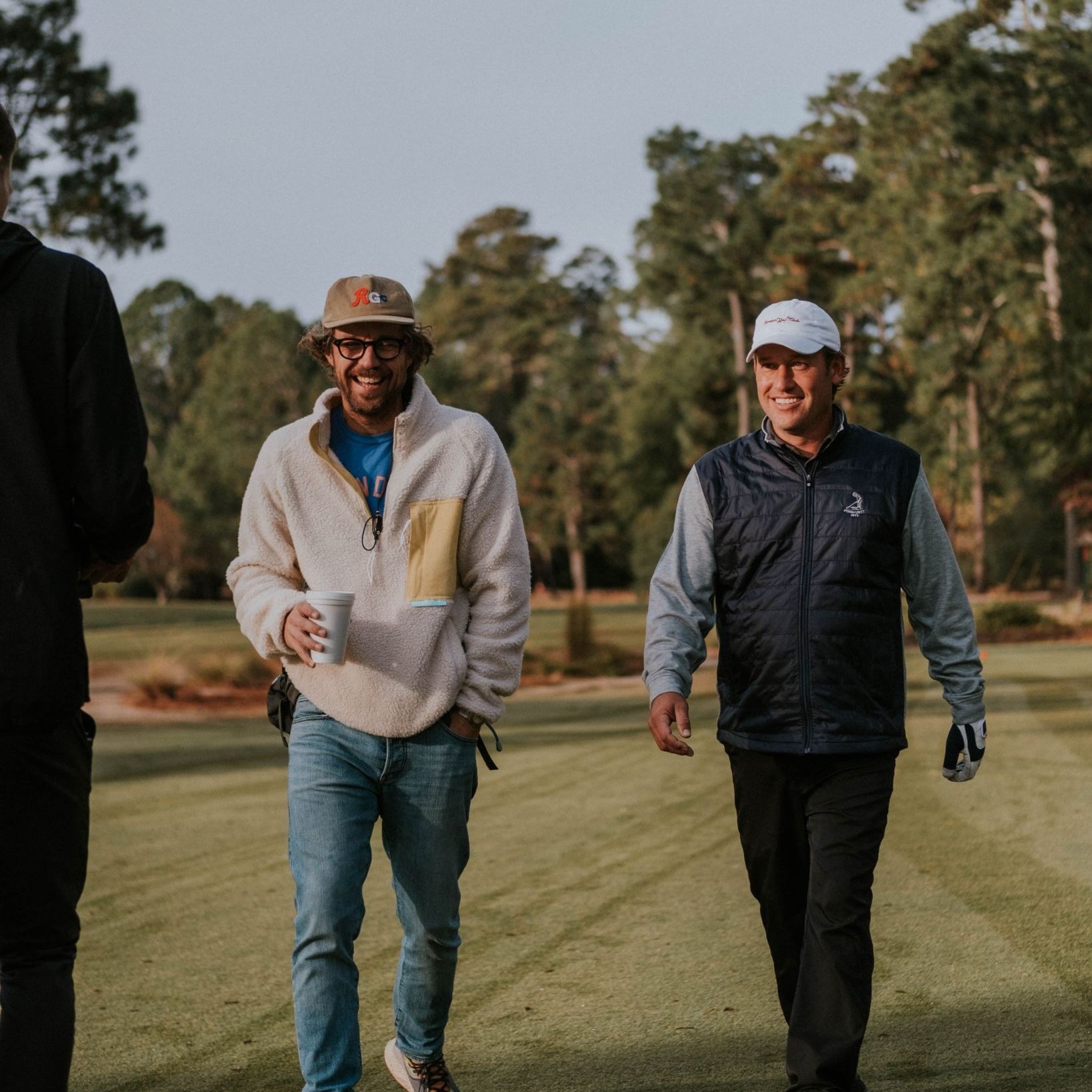 Two smiling men walking along a pine-lined fairway, one holding a coffee cup