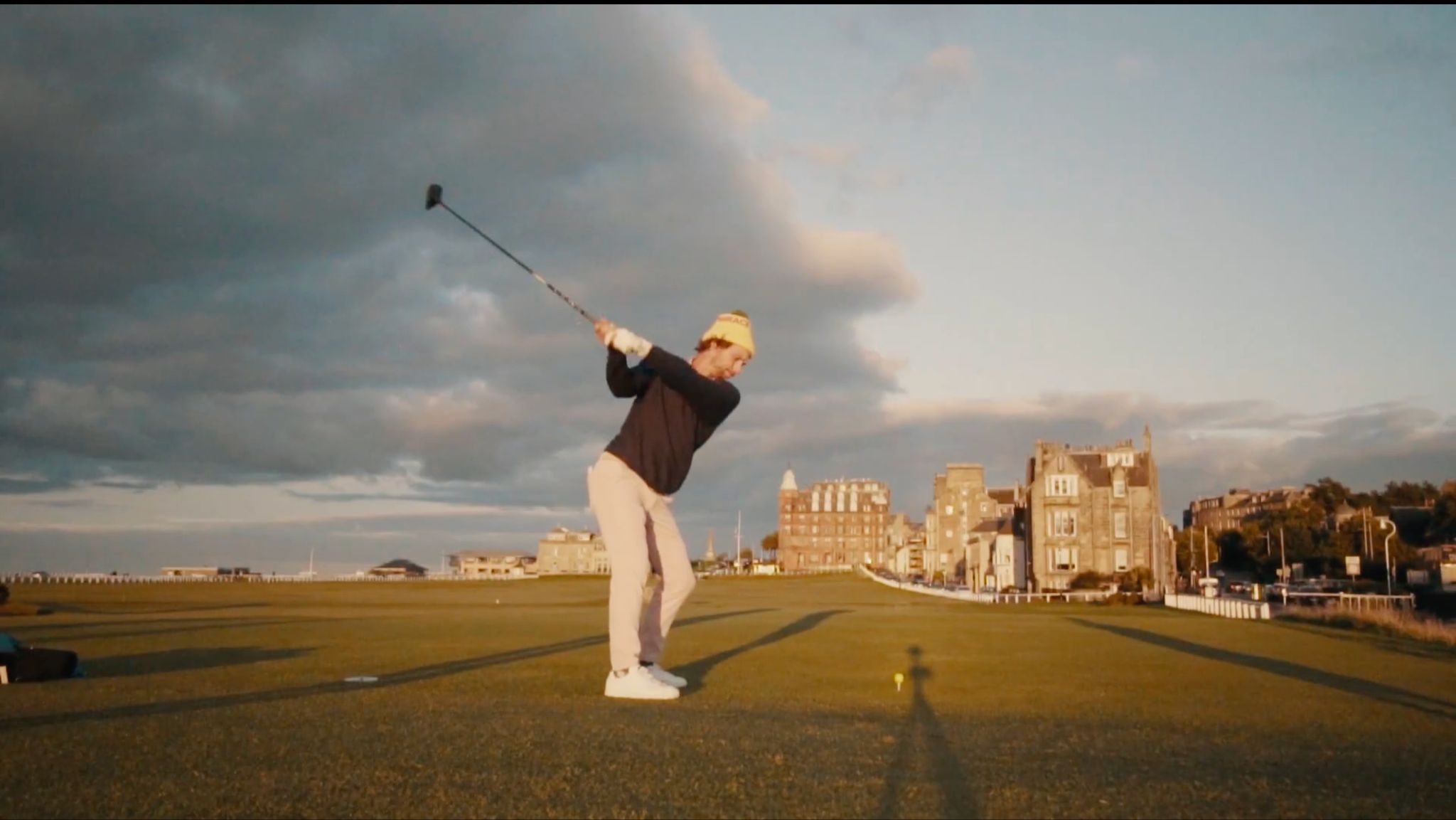 Golfer mid-swing on a links course at sunset beside historic stone buildings