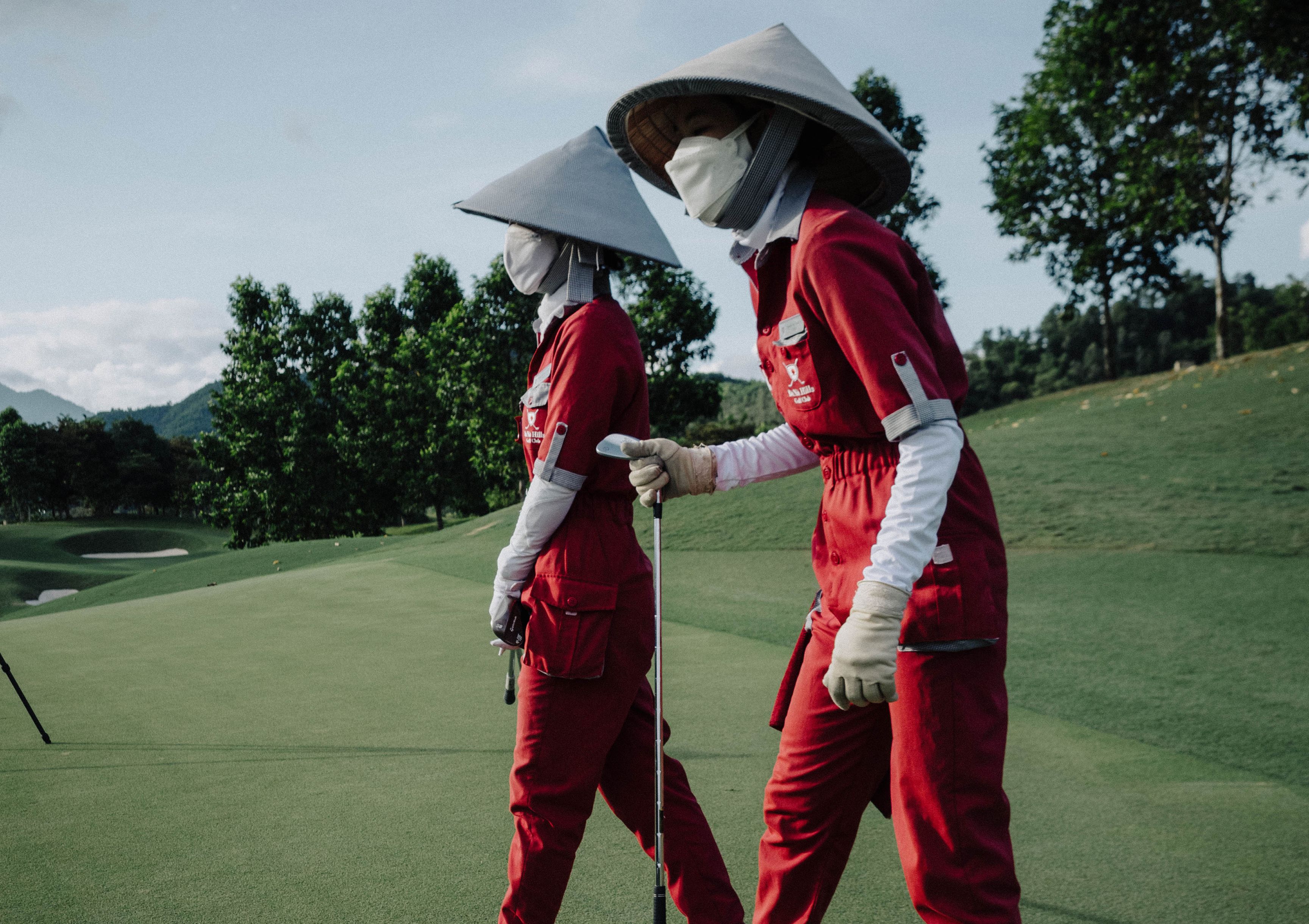 Two caddies in red jumpsuits and conical hats walking a golf course, one holding a club