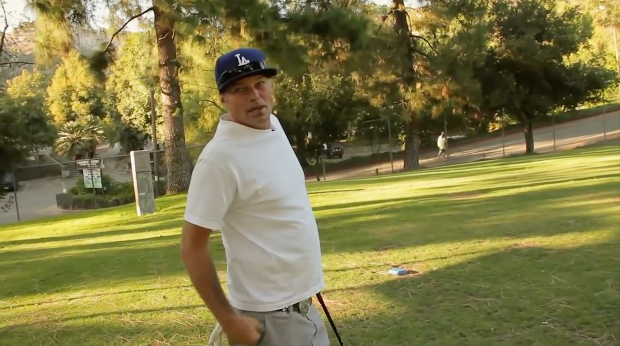 Man in a white tee and LA cap holding a golf club on a sunny park lawn