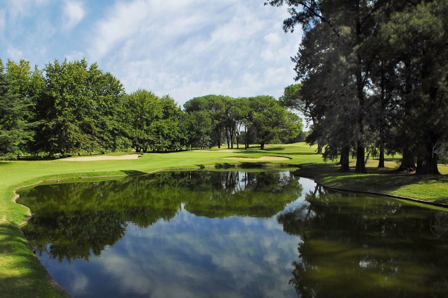 Tree-lined parkland golf hole with a still pond reflecting the sky