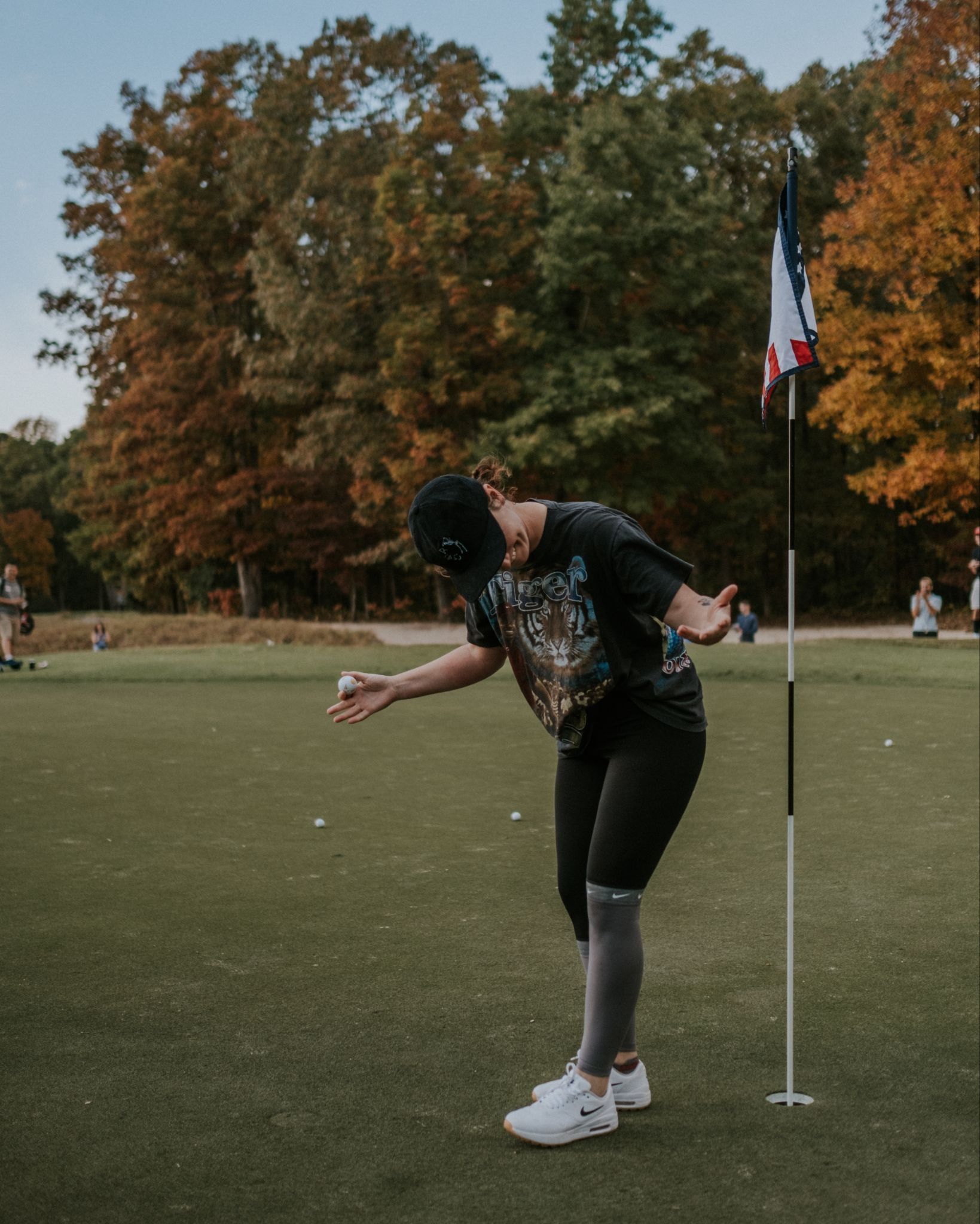 Golfer bowing beside the flagstick holding a ball, with autumn trees behind