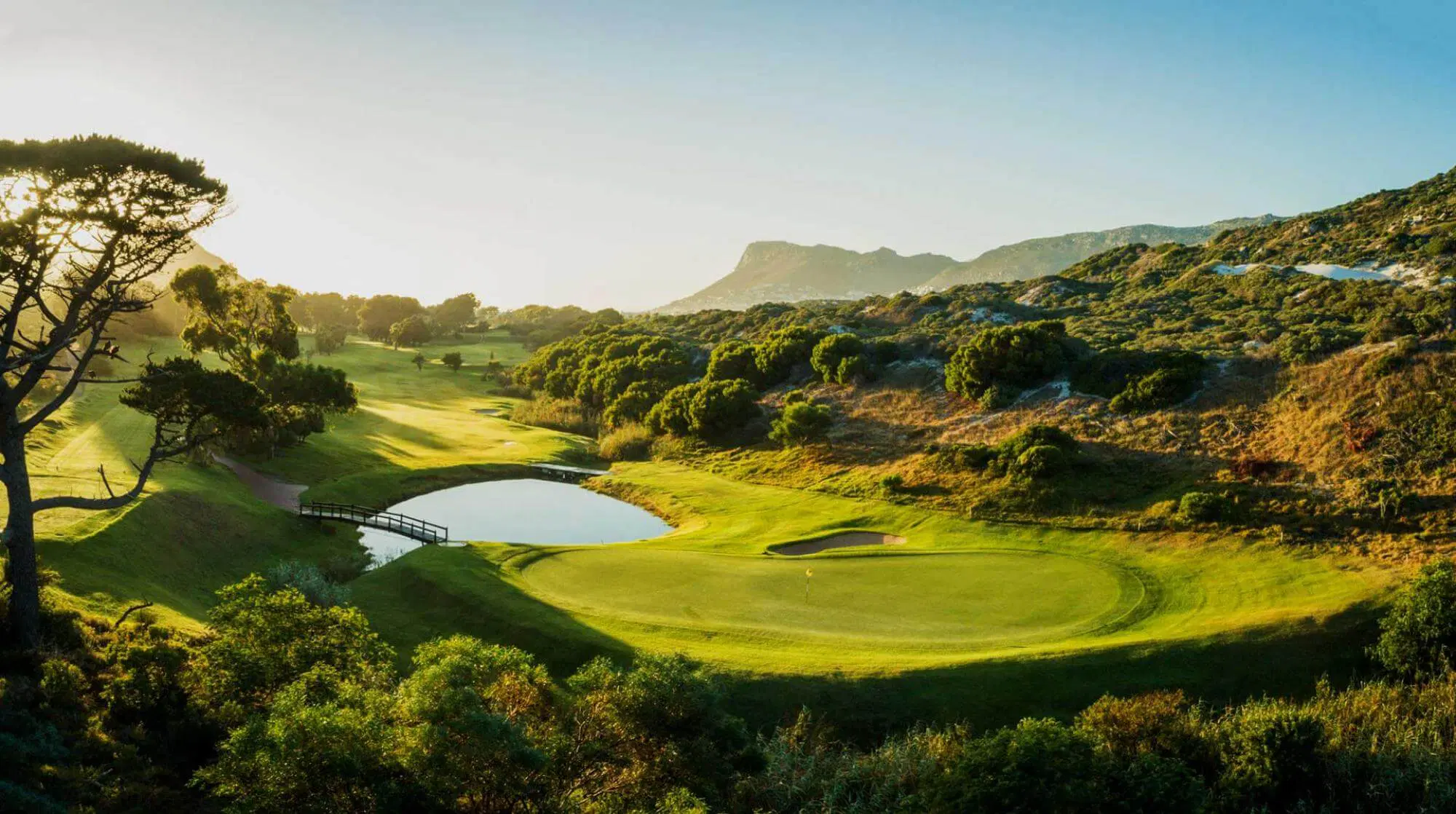 Golf green beside a pond and footbridge among hills at sunset