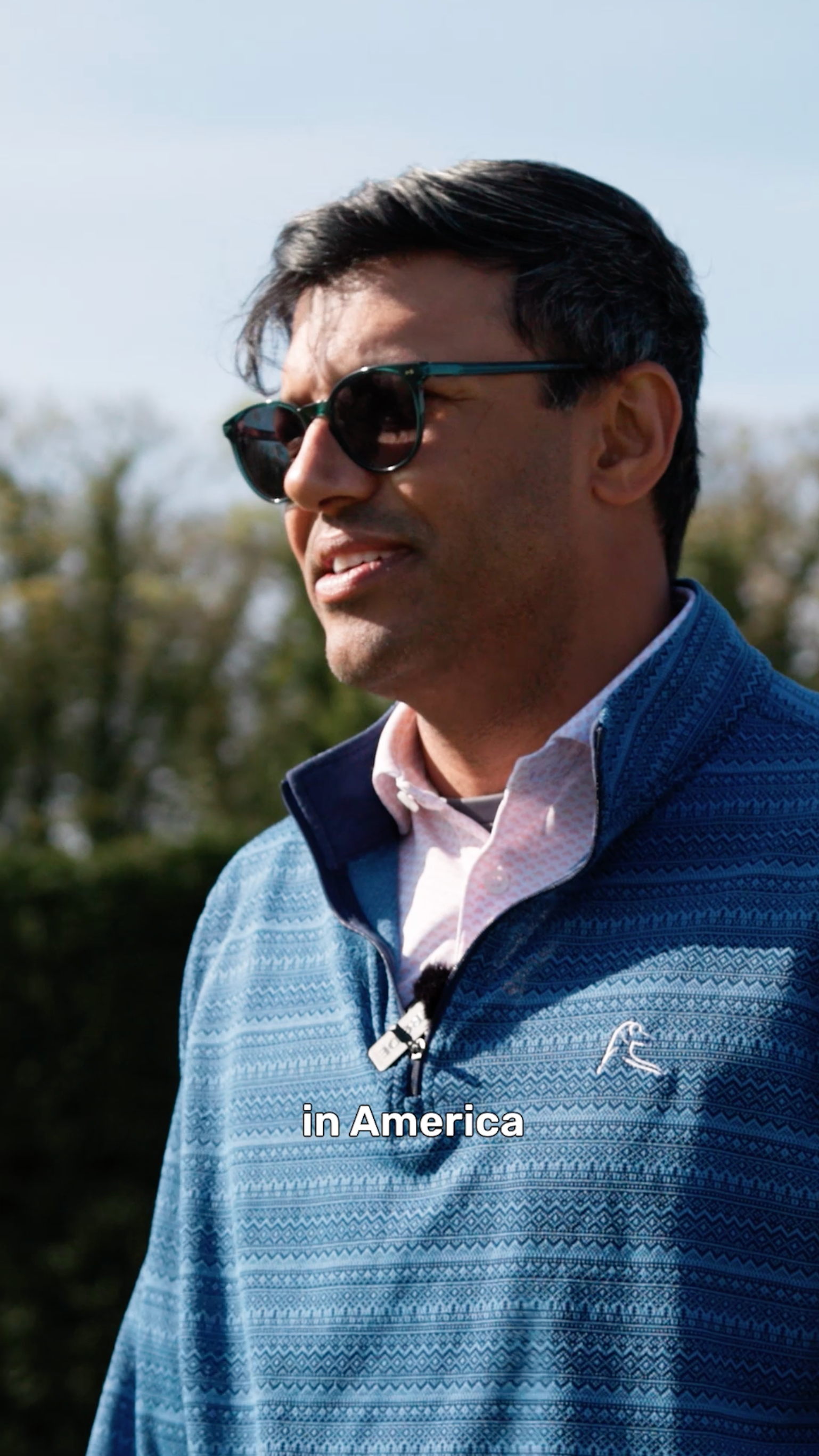 Man in sunglasses and a blue quarter-zip speaking outdoors, captioned "in America"