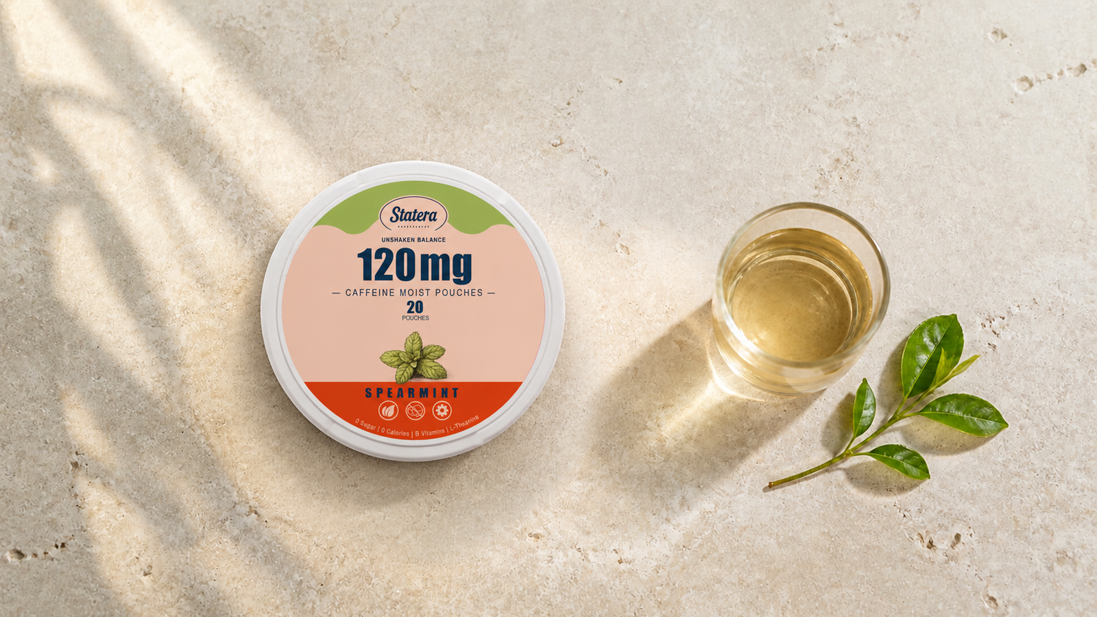 STATERA Spearmint 120mg caffeine pouch can beside a clear glass and a small tea-leaf sprig on a warm stone surface