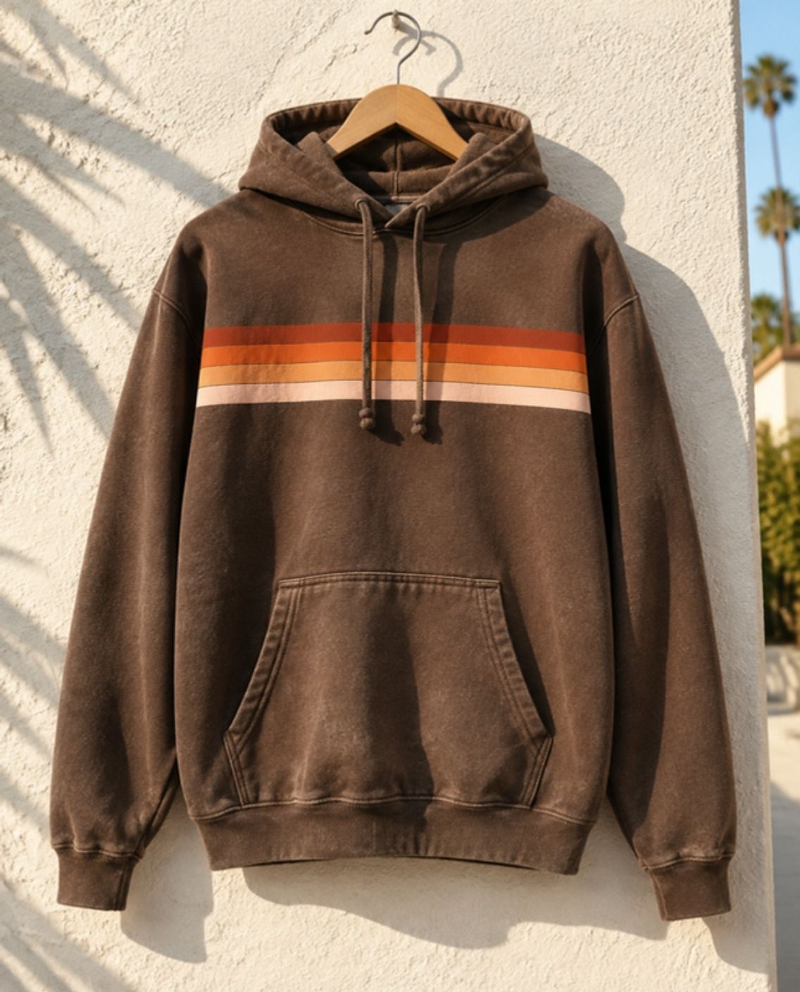 Sun-faded brown hoodie with a retro stripe across the chest, hanging on a stucco wall
