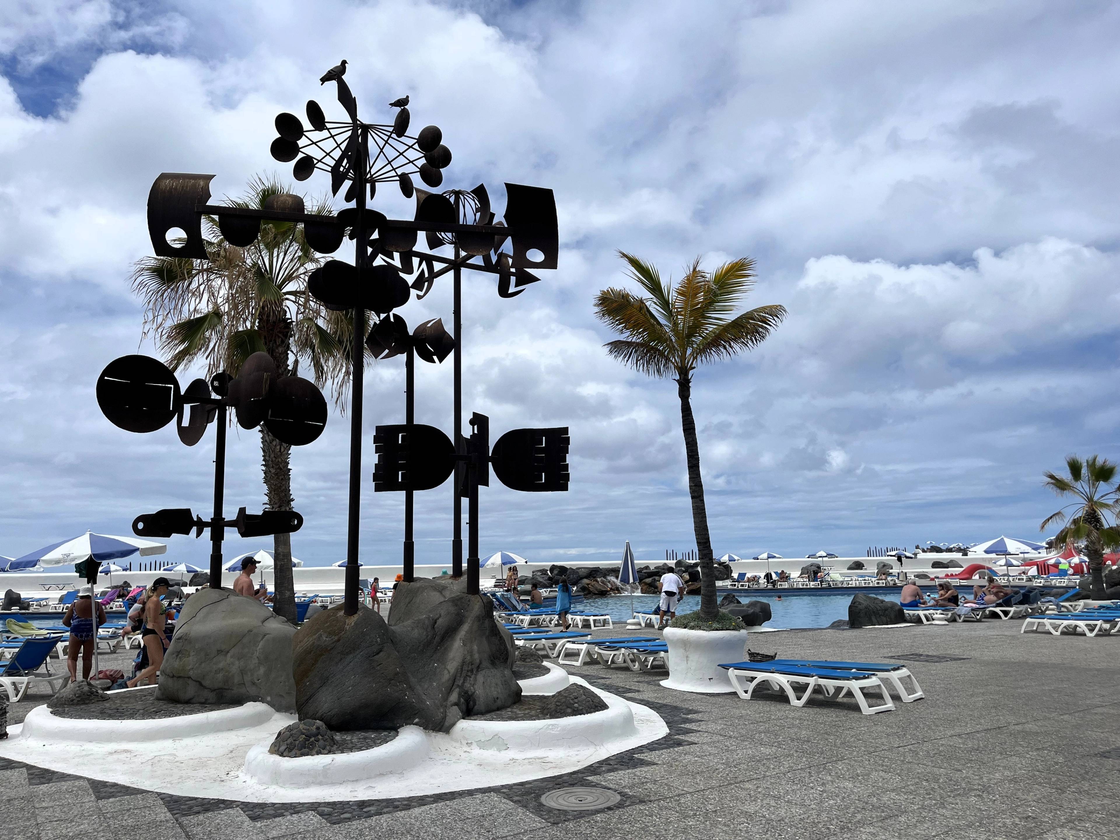 Nowoczesna rzeźba składająca się z geometrycznych elementów, obok palma, a w tle baseny i leżaki