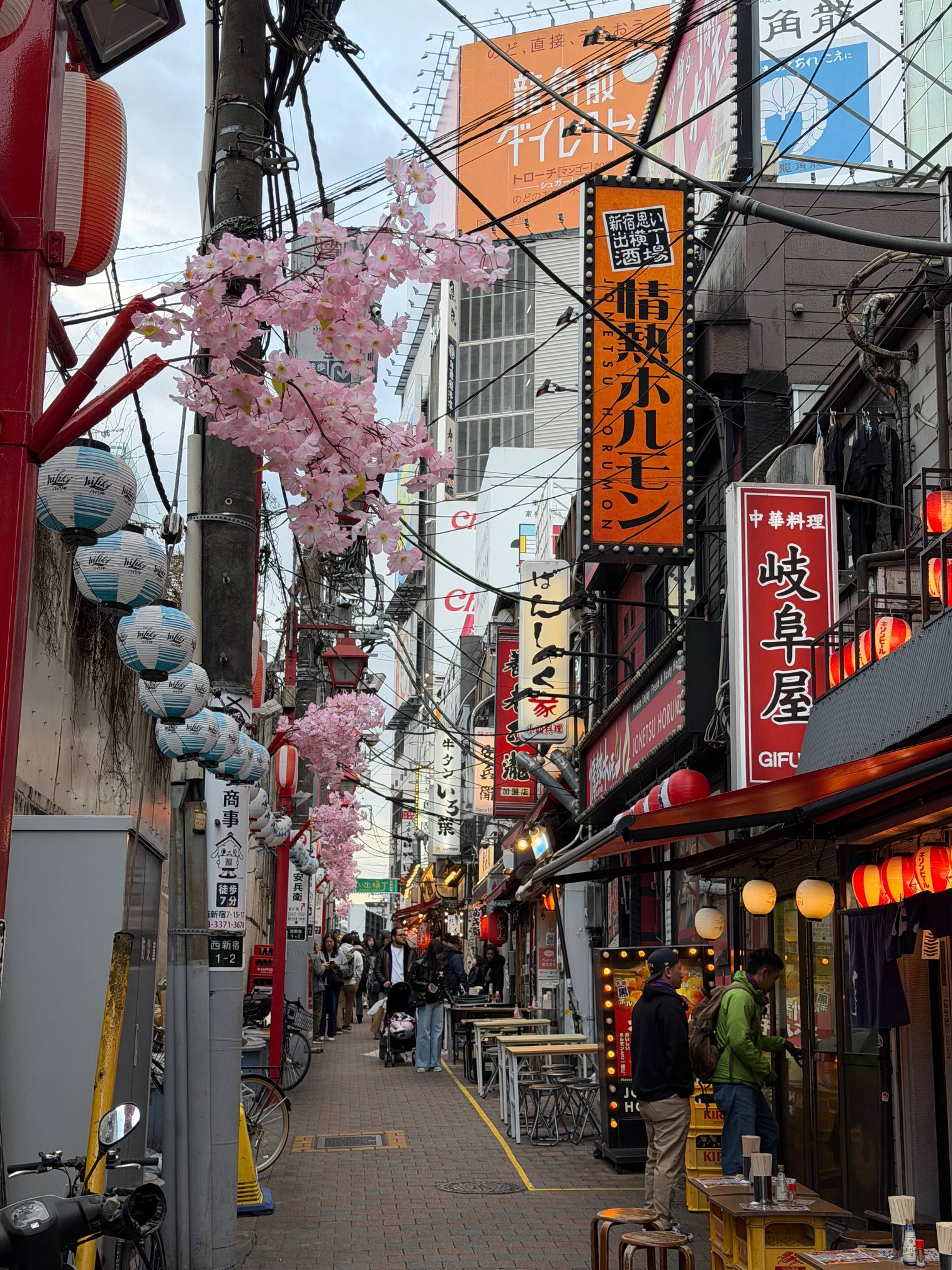 Wąska ulica z restauracjami, wiszącymi papierowymi lampionami i różowymi kwiatami wiśni, przy której stoją stoliki i przechodnie.