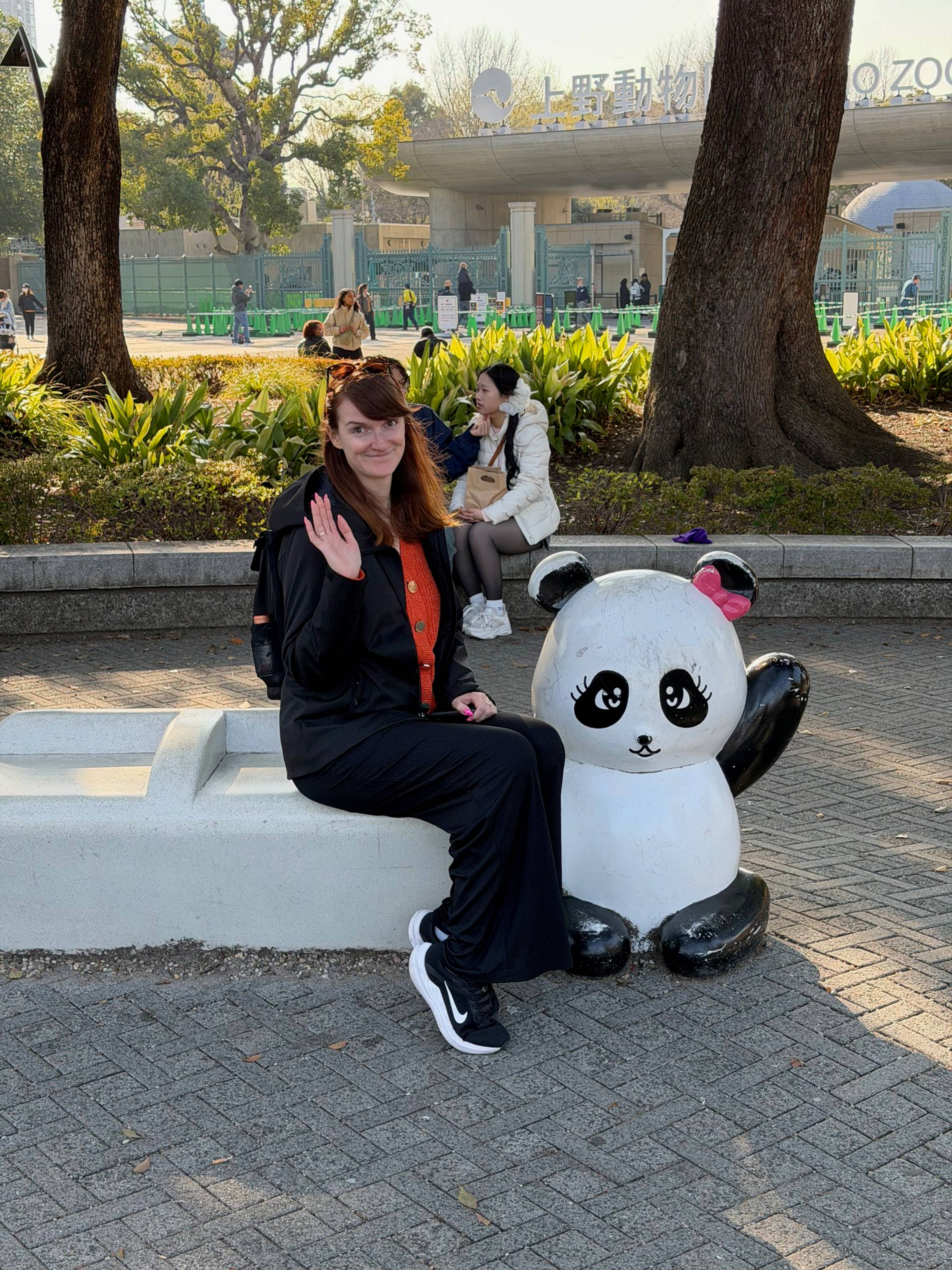 Osoba siedzi na betonowej ławce obok figurki pandy w ogrodzie zoologicznym Ueno w Tokio, a w tle widoczna jest brama zoo, drzewa i przechodnie.