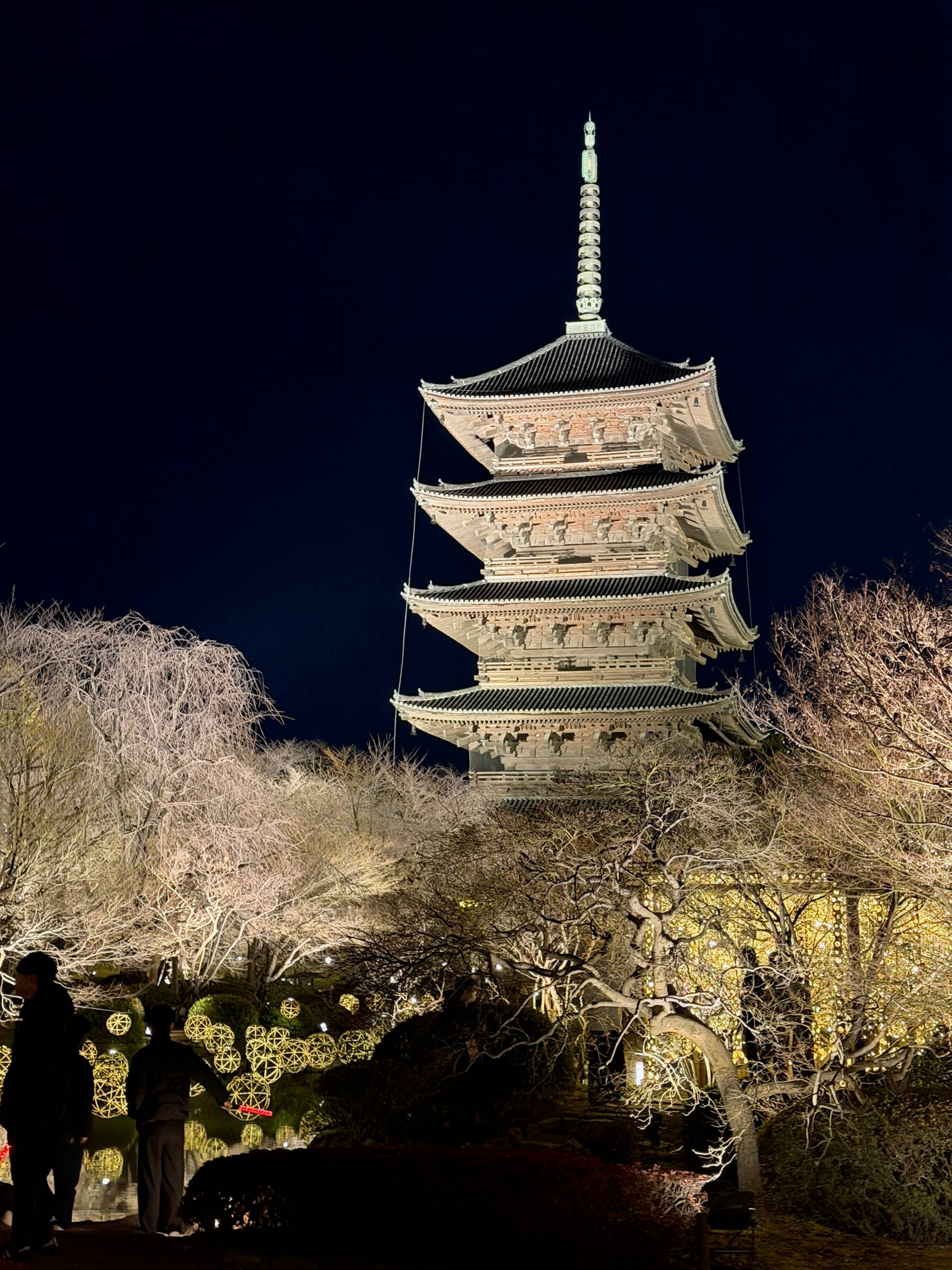 Wielopiętrowa japońska pagoda jest oświetlona nocą i otoczona drzewami, a na pierwszym planie stoją odwiedzający na alejkach ogrodu.