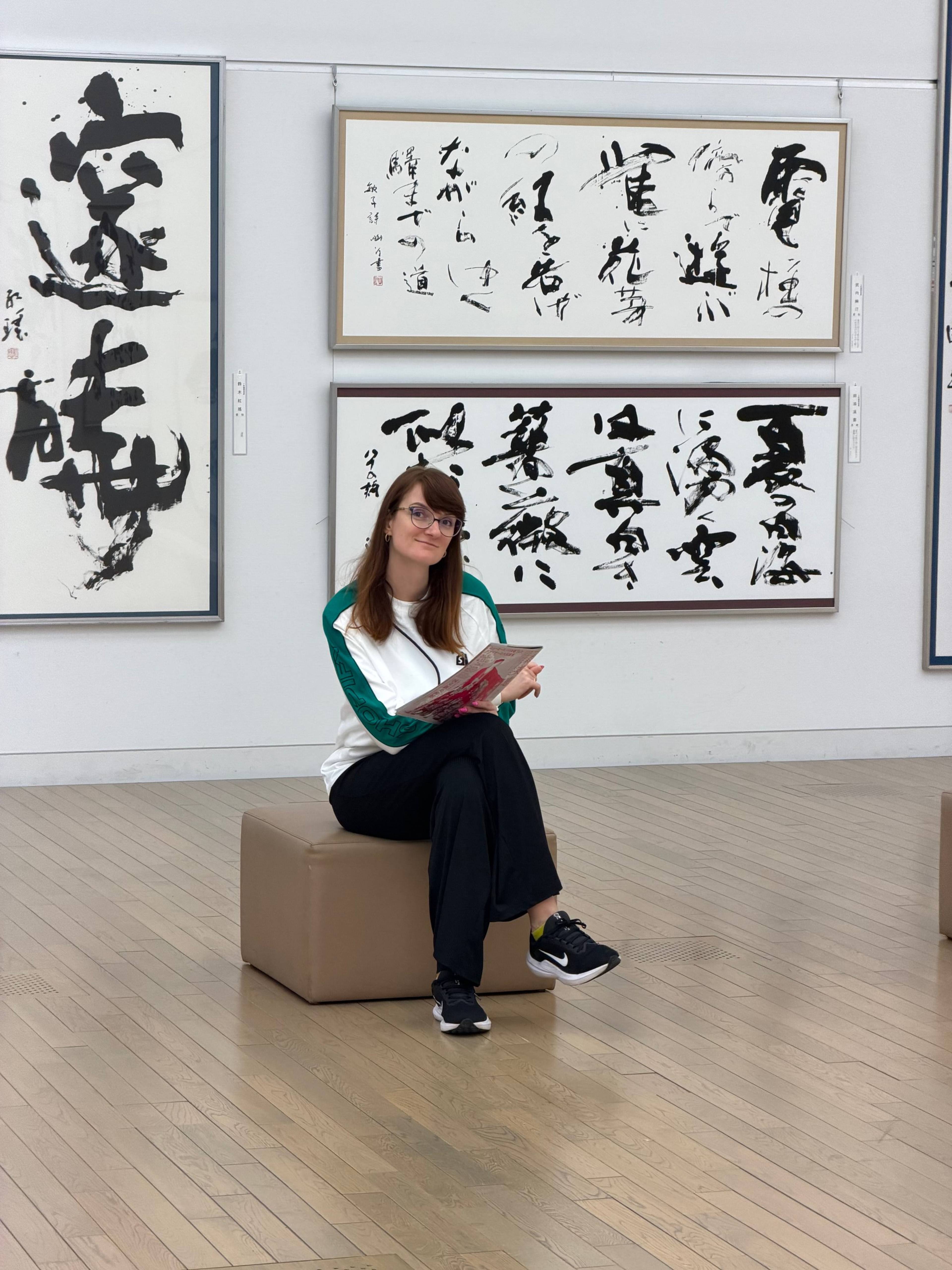 Osoba siedzi na niskim siedzisku w galerii sztuki, trzyma książkę, a na ścianach za nią wiszą duże prace z japońską kaligrafią.