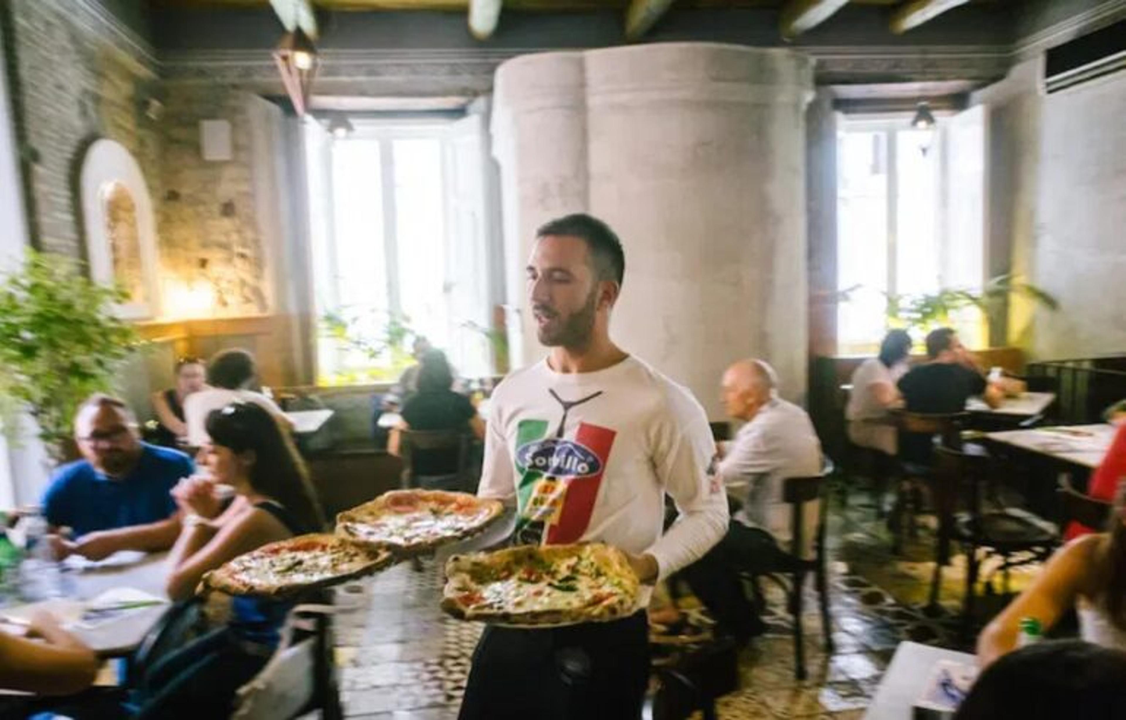 Kelner niesie trzy pizze, wokół stoliki i goście, restauracja