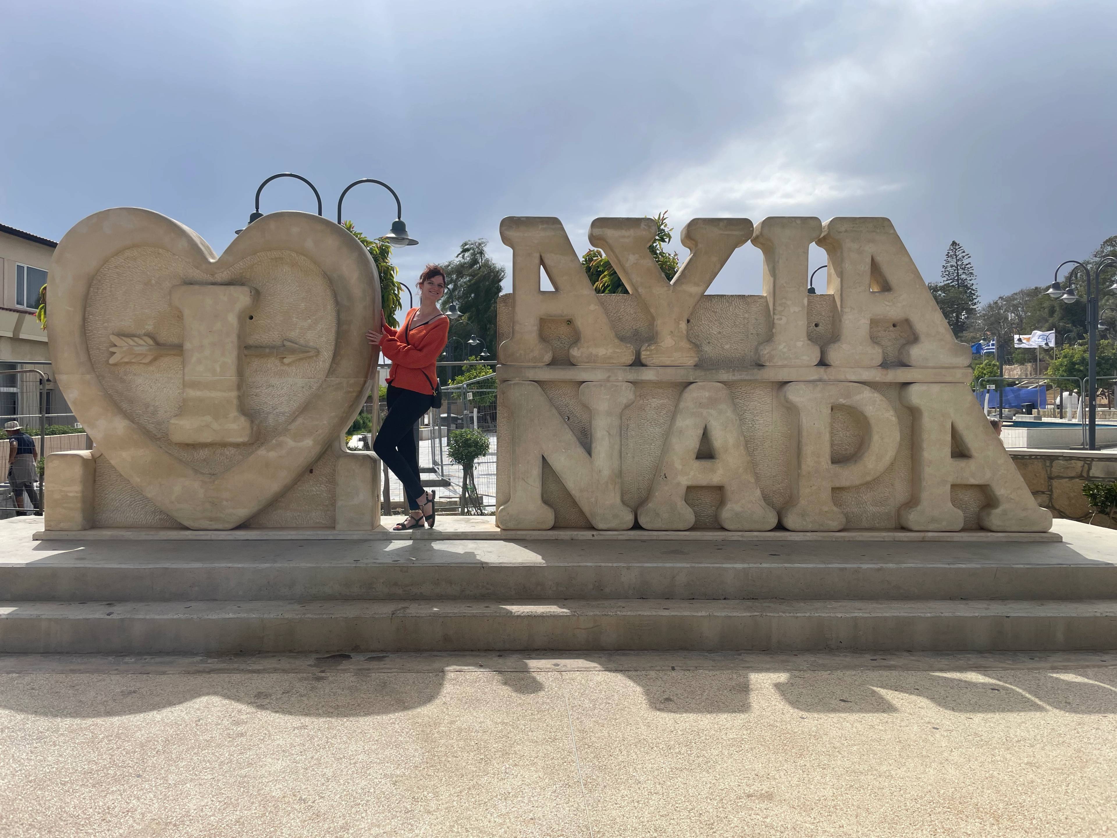 Kamienny napis ‘I ❤️ Ayia Napa’ w centrum miasta na Cyprze, ustawiony na placu z latarniami w tle.