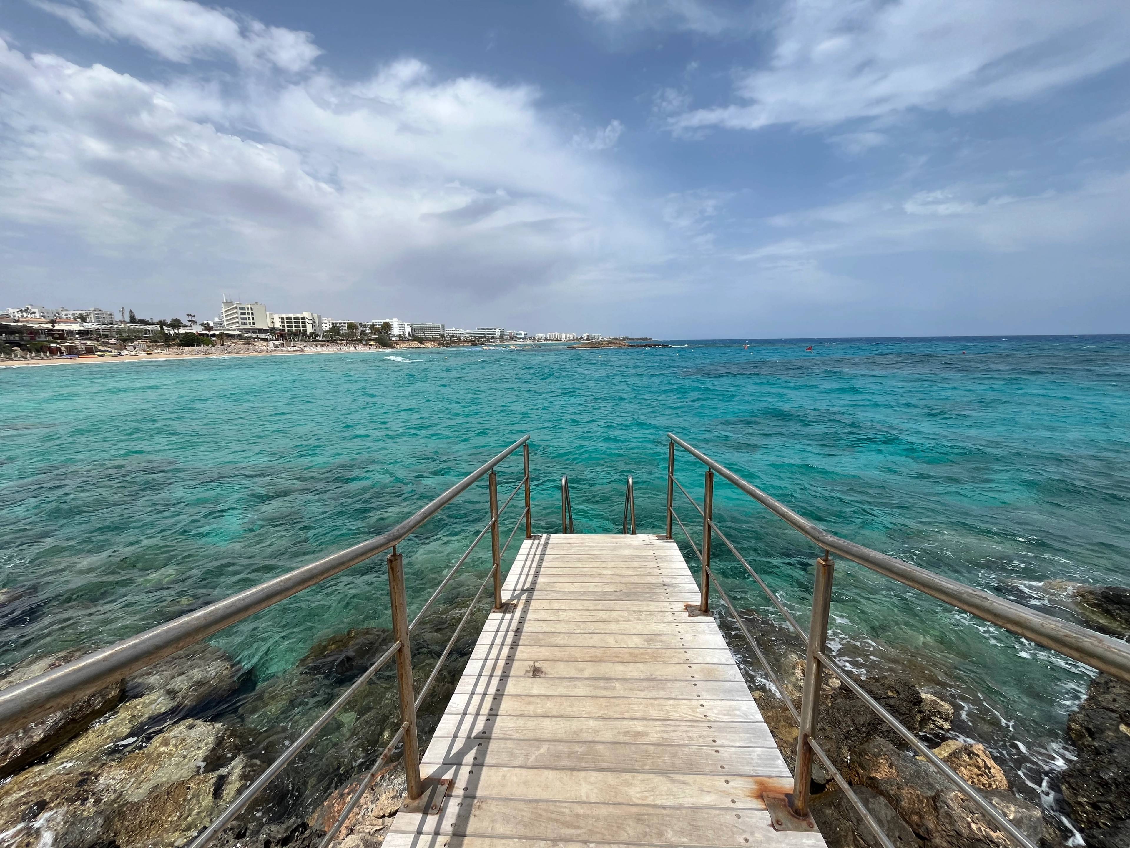 Drewniany pomost z metalowymi poręczami wychodzący na turkusowe morze w Protaras na Cyprze.